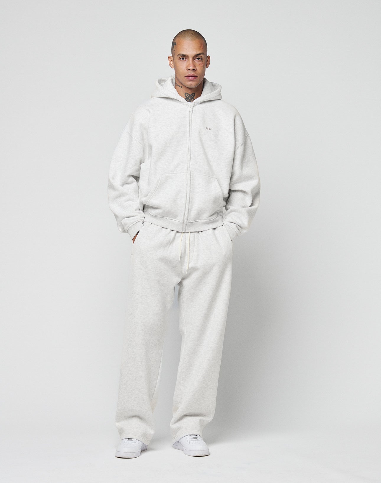 A person stands facing forward against a white background, wearing the LIVE FAST DIE YOUNG Basic Hooded Zip and matching sweatpants with logo print, plus white sneakers. Their hands are in their pockets.