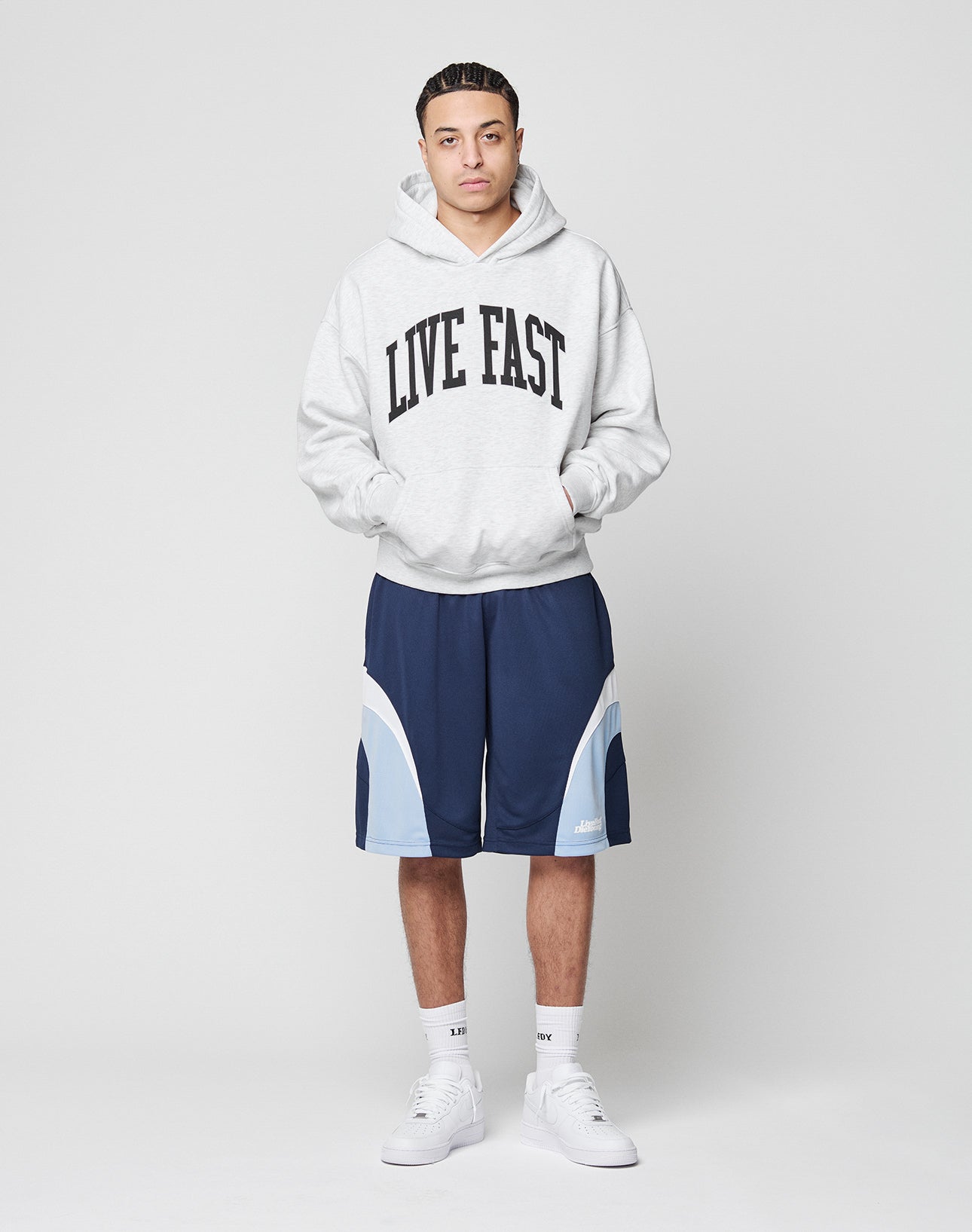 A person stands facing forward, wearing the LIVE FAST DIE YOUNG Live Fast College Hooded in light gray with bold black front print, paired with navy and light blue athletic shorts, white socks, and white sneakers against a plain background.