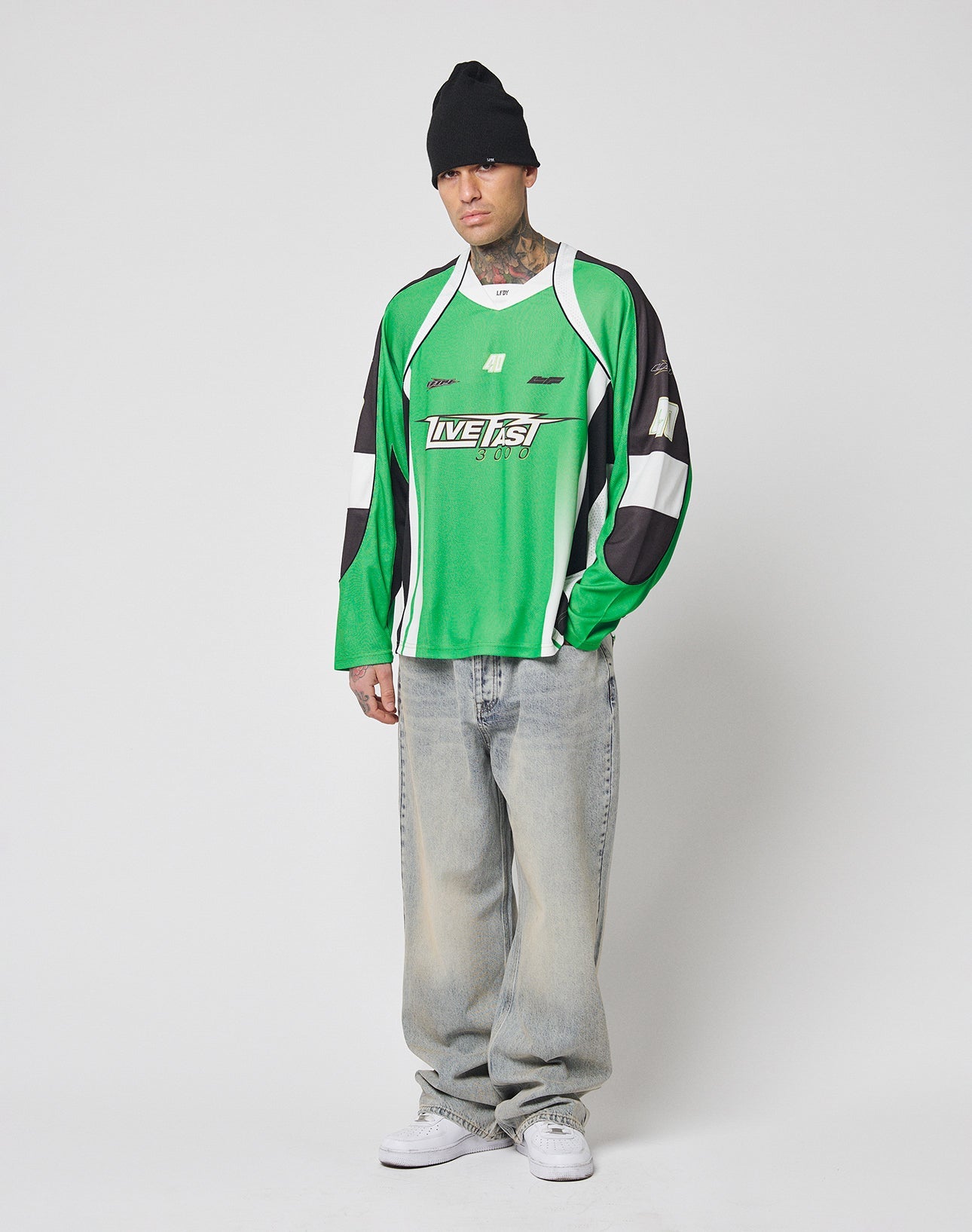 A man wears the LIVE FAST DIE YOUNG LFDY Motorcross Jersey, a green oversize-fit sports top with multi-logo prints, paired with a black beanie, loose faded jeans, and white sneakers against a plain backdrop.