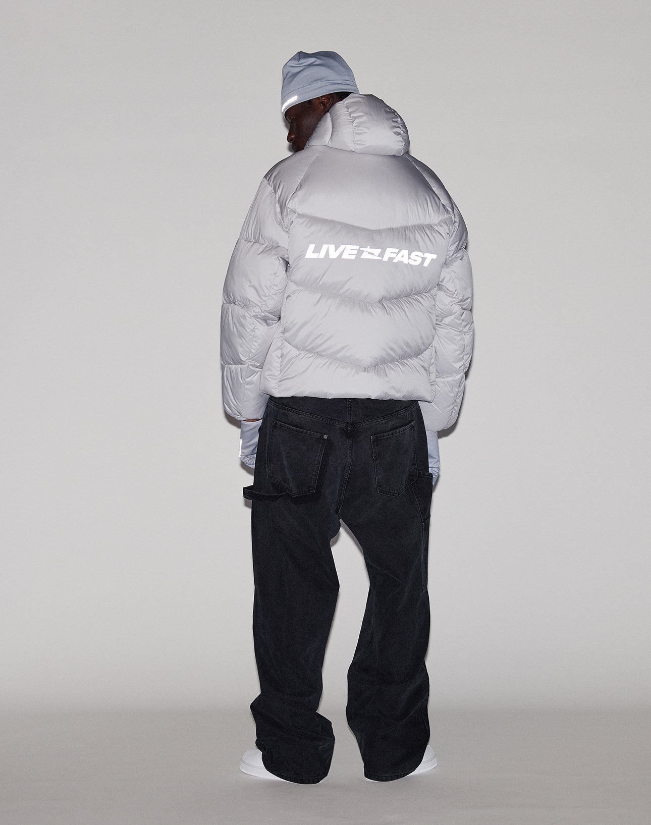 A person faces away, wearing the LIVE FAST DIE YOUNG Live Fast Puffer Jacket in oversize light gray with reflective "LIVE FAST" print on the back, black jeans, white shoes, and a matching beanie, set against a plain light background.