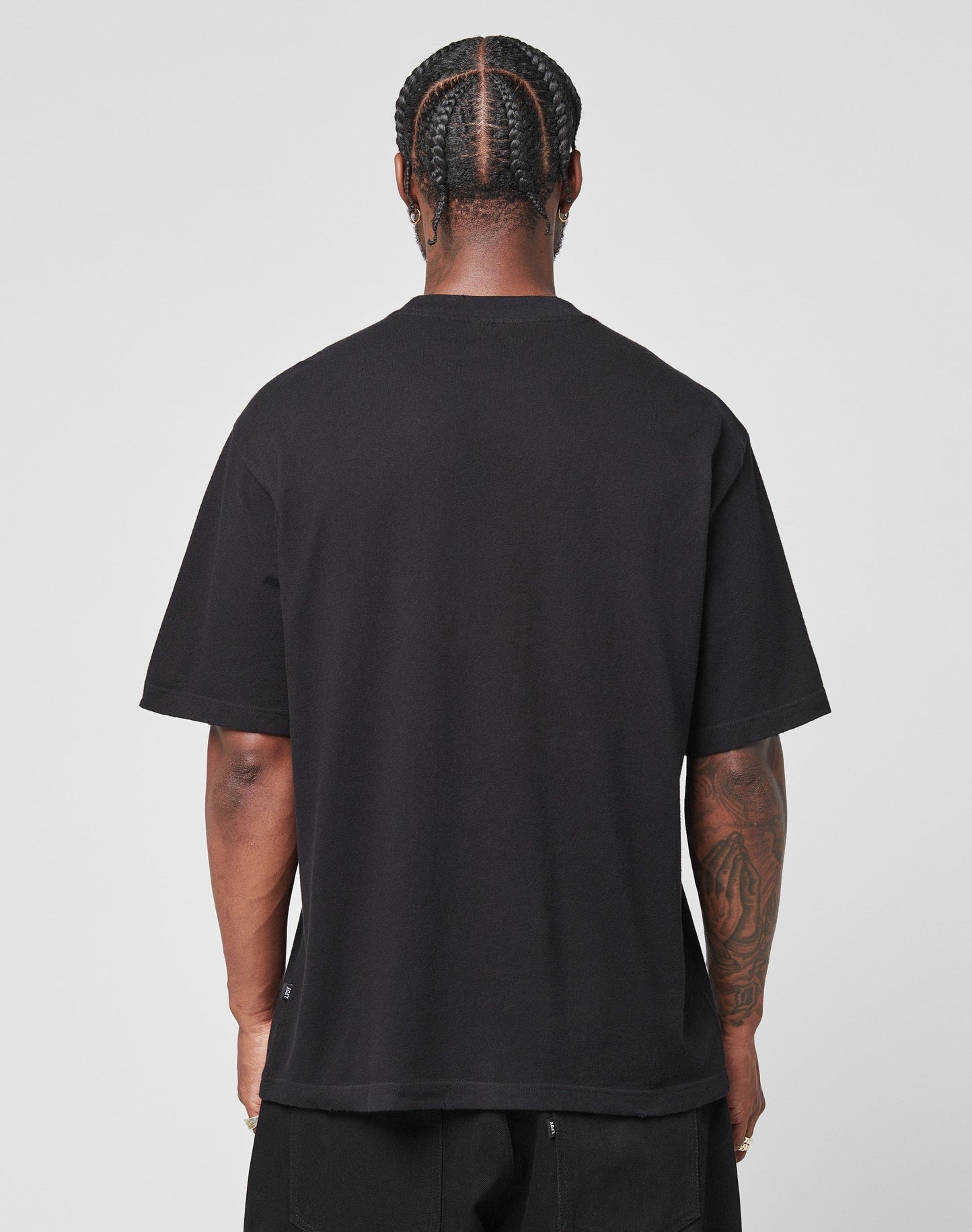 A person with braided hair and arm tattoos faces away from the camera, wearing the LIVE FAST DIE YOUNG LFDY Vintage Tee—an oversized black shirt with distressed edges—and black pants against a plain light background.