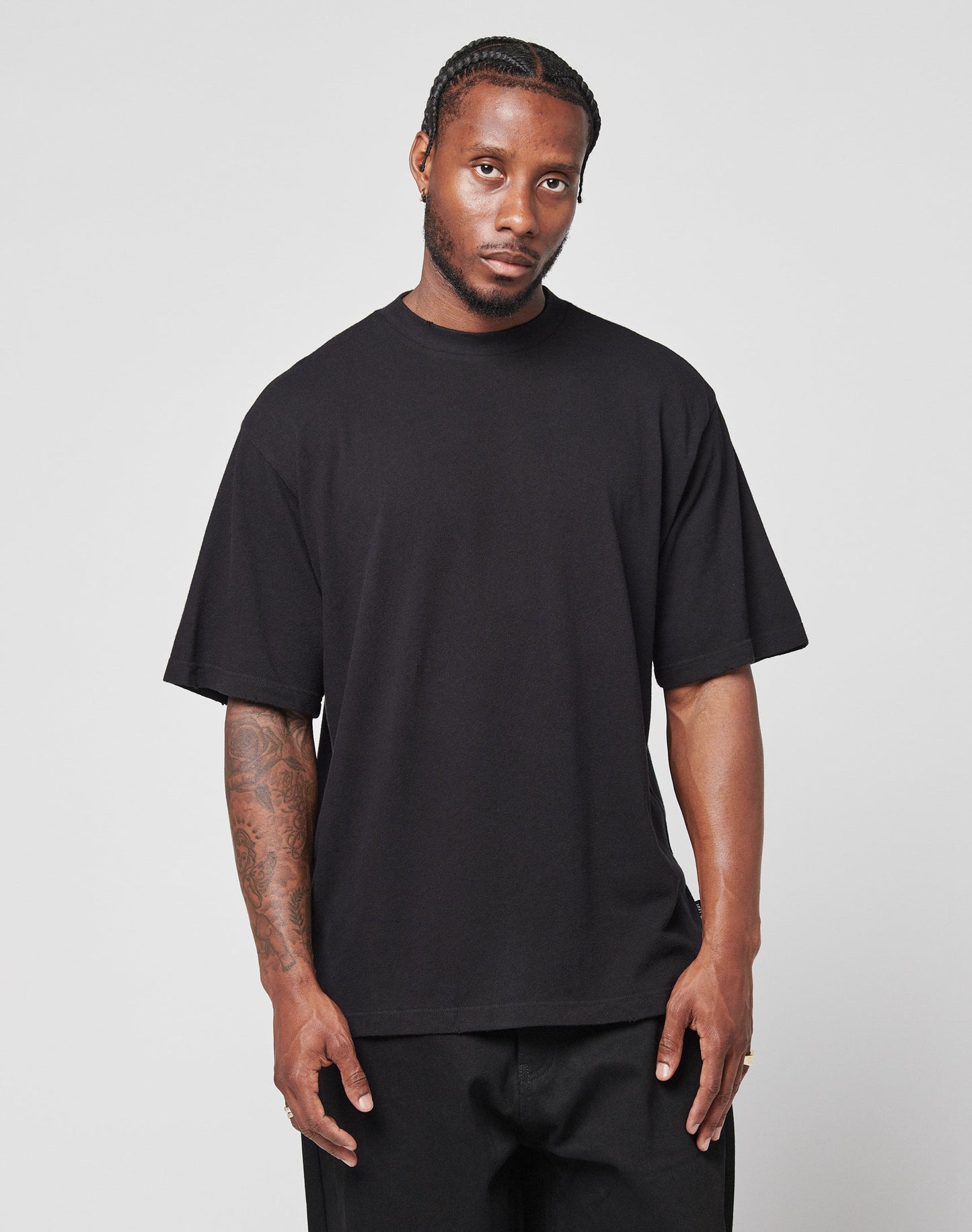 A man with braided hair and forearm tattoos stands against a plain background, wearing the LIVE FAST DIE YOUNG LFDY Vintage Tee in an oversize fit with distressed edges and black pants, looking at the camera with a neutral expression.