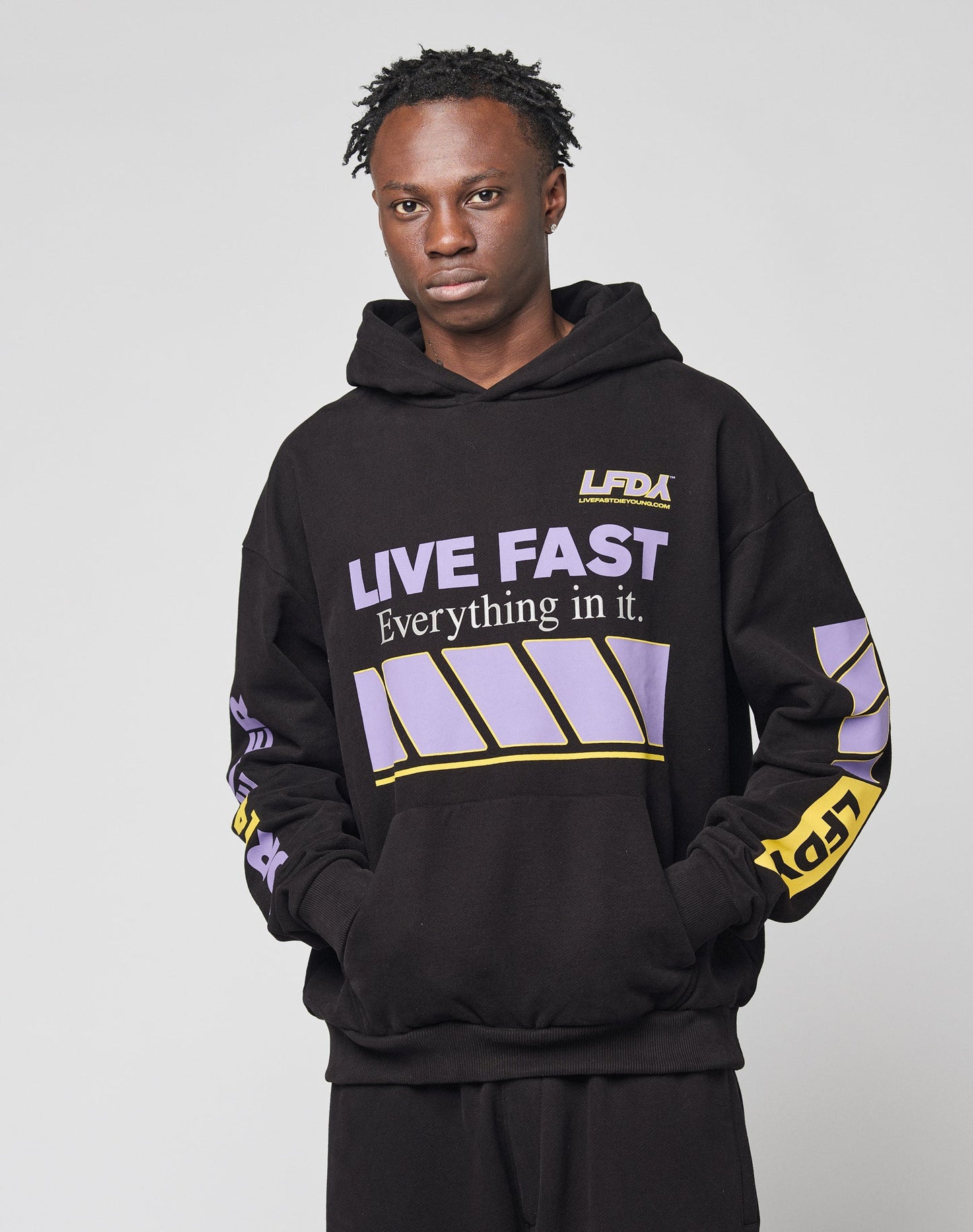 A person wears the LIVE FAST DIE YOUNG LFDY Racing Hooded black hoodie with glossy purple and yellow graphics that say LIVE FAST Everything in it, paired with matching black pants, against a plain light background.