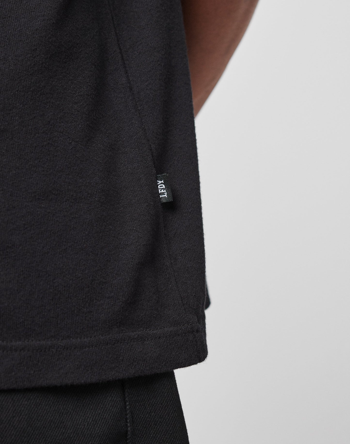 Close-up of the lower side of an oversize fit black LFDY Vintage Tee by LIVE FAST DIE YOUNG, featuring a small black fabric tag sewn into the side seam, paired with black pants.