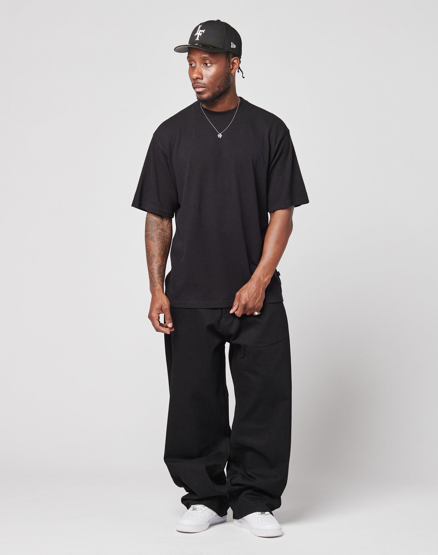 A man stands against a plain background wearing a black LFDY Vintage Tee by LIVE FAST DIE YOUNG with distressed edges, paired with loose black pants, a black baseball cap, and white sneakers, his hands relaxed by his sides.