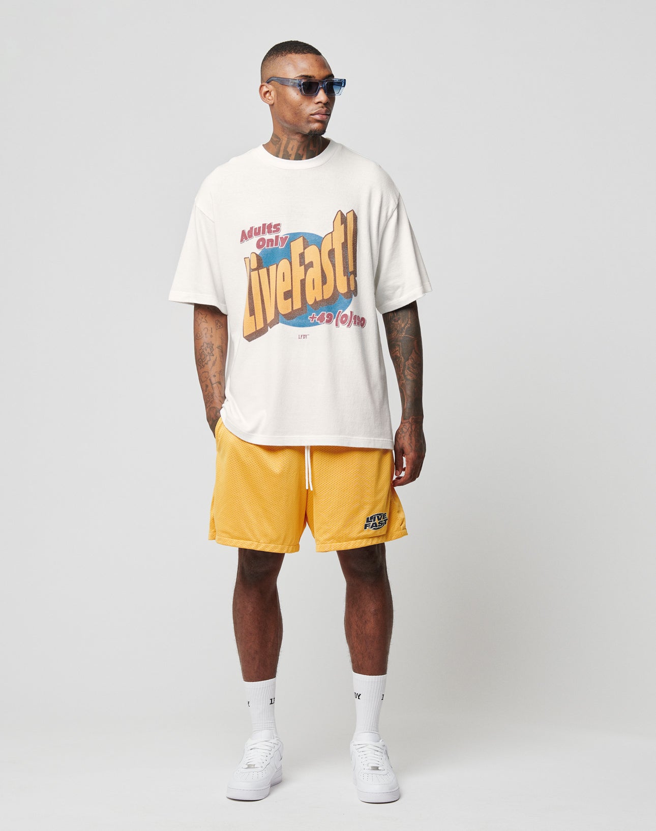 A man in sunglasses, yellow shorts, white crew socks and sneakers wears a LIVE FAST DIE YOUNG LFDY Adults Only Tee, standing against a plain light gray background.