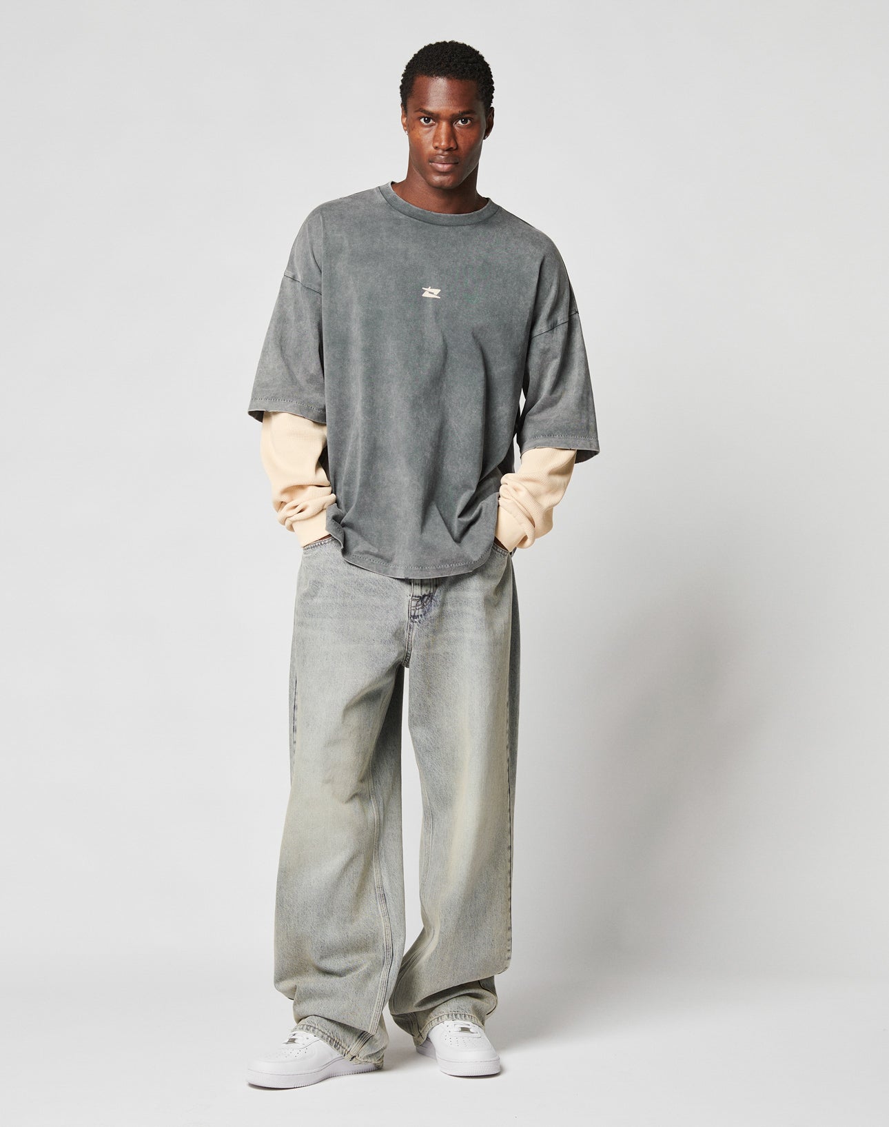 A man stands facing forward against a light gray background, wearing the LIVE FAST DIE YOUNG LFDY Double Layered Longsleeve, baggy faded jeans, and white sneakers, with his hands in his pockets.