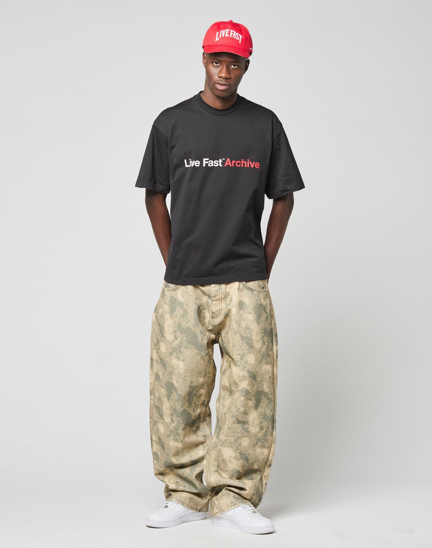 A man stands against a plain background wearing a red Live Fast cap, LIVE FAST DIE YOUNG Live Fast Archive Tee with front print and oversized fit, loose camouflage pants, and white sneakers. His hands are behind his back.