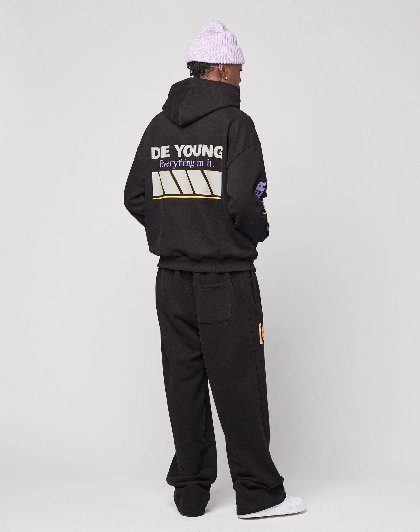 Wearing an oversize black LFDY Racing Hooded by LIVE FAST DIE YOUNG with glossy DIE YOUNG, Everything in it. print on the back, black pants, a lavender beanie, and white shoes, a person stands facing away from the camera against a plain background.