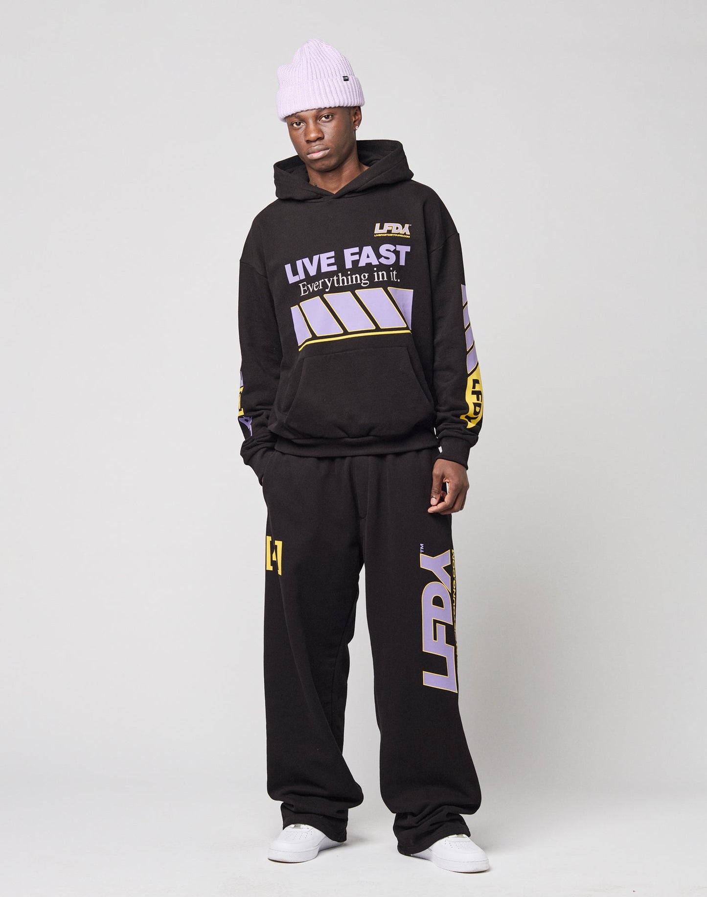 A person wears a lavender beanie, black LIVE FAST DIE YOUNG hoodie with graphic designs, LFDY Racing Wide Jogger sweatpants with LFDA text and prints on the legs, and white sneakers against a plain light gray background.