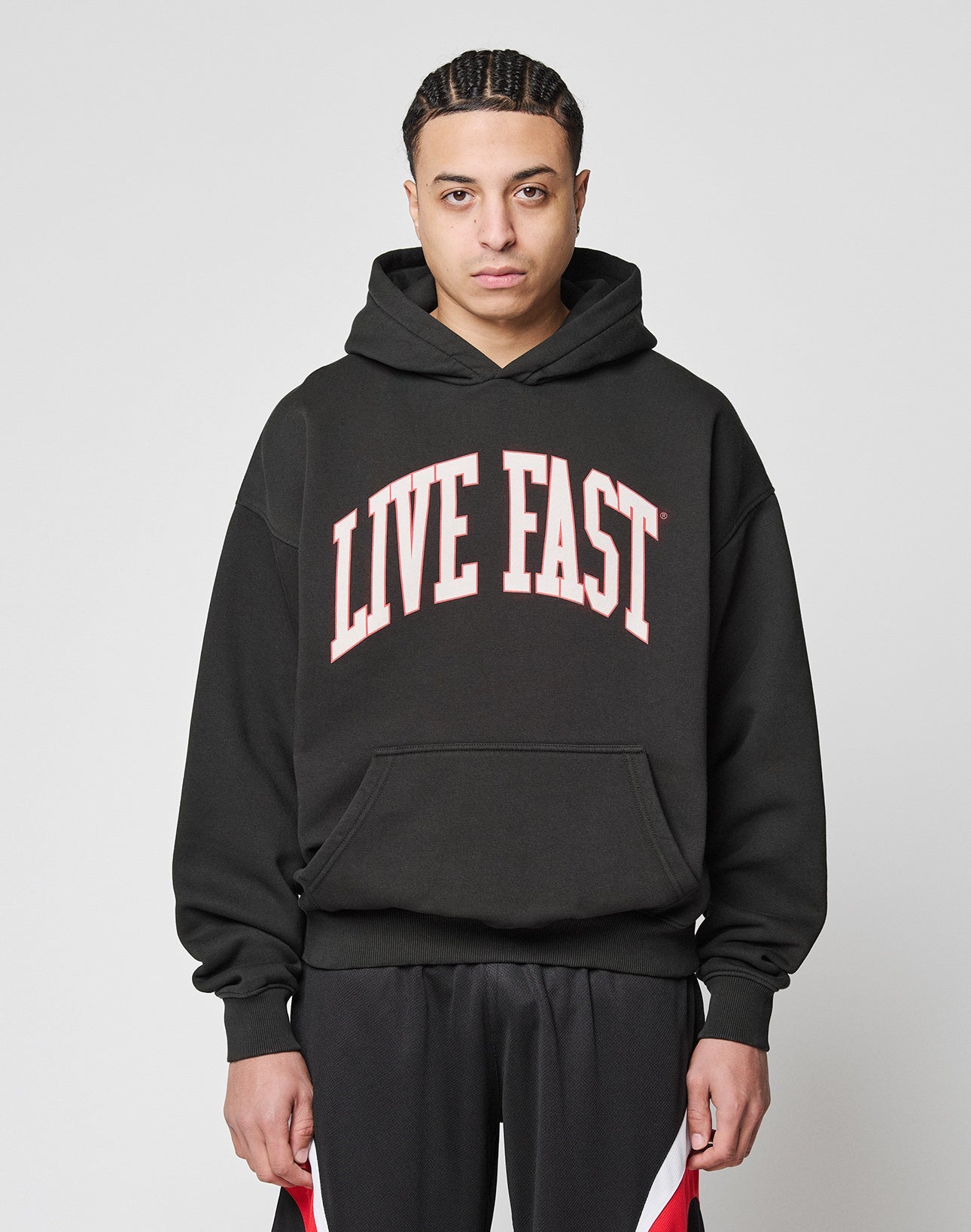 A person with braided hair models the LIVE FAST DIE YOUNG Live Fast College Hooded—an oversize black hoodie with a bold white and red front print, made from 65% Baumwolle, against a plain light background.
