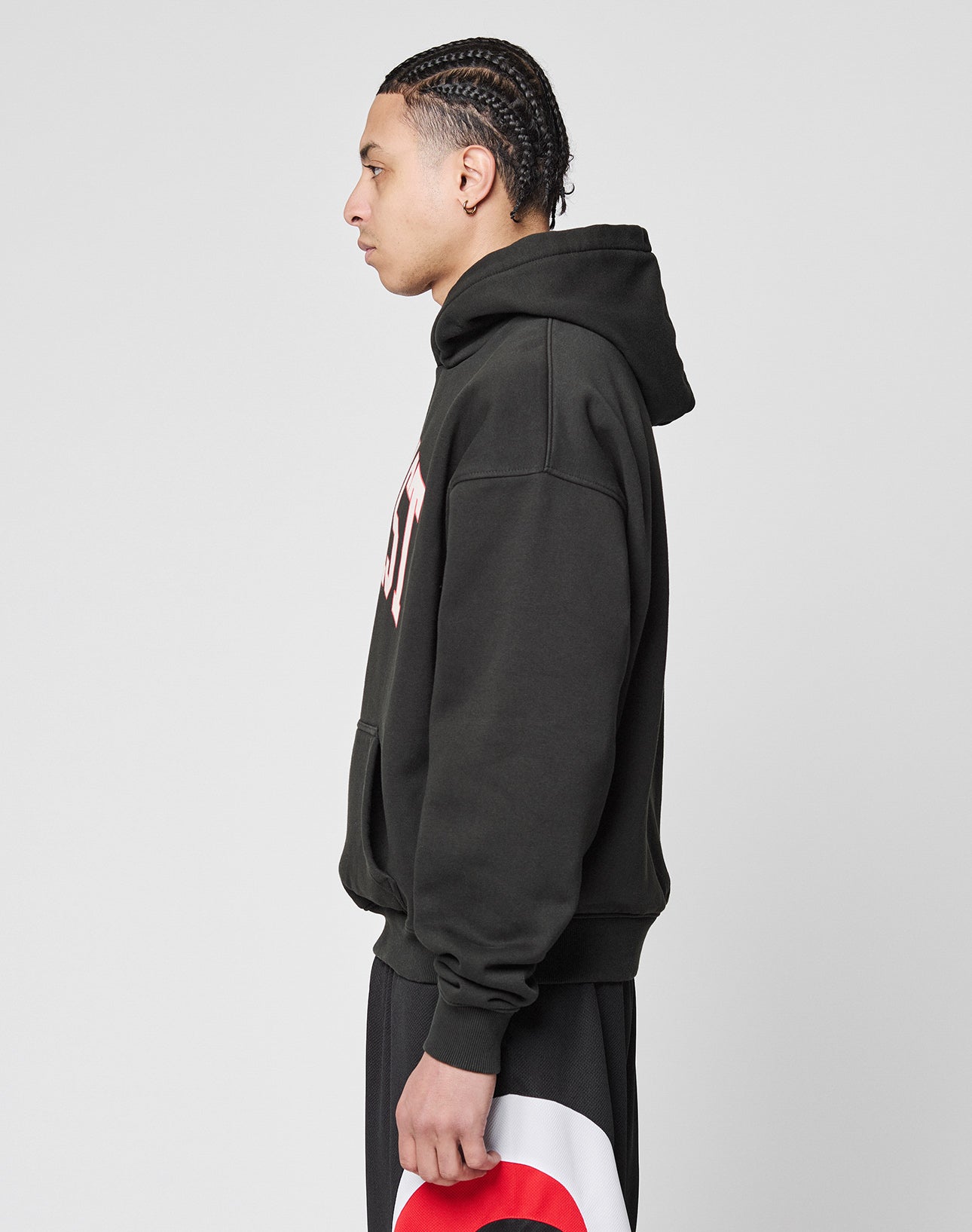 A young person with braided hair wears the LIVE FAST DIE YOUNG Live Fast College Hooded in black with a white front print and matching black pants with a bold red and white circular design, all in an oversize fit, against a plain light gray background.