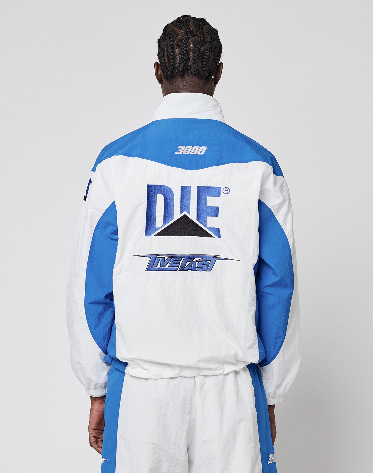A person with braided hair, seen from behind, wears the LFDY Trackjacket 3000 by LIVE FAST DIE YOUNG. This blue and white jacket features a Multilogo Stick, bold DIE and LIVE FAST lettering, a black triangle graphic, and a Reißverschluss-Puller detail.