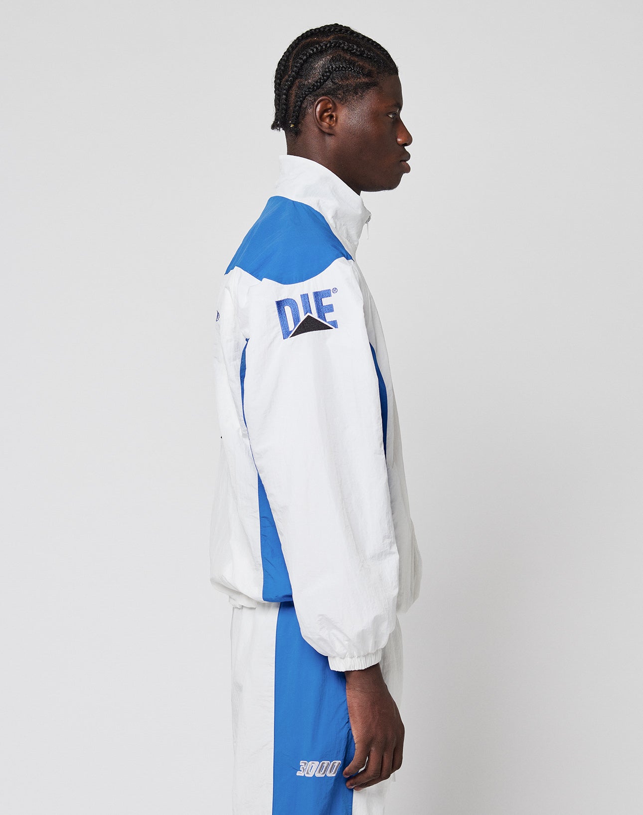 A person with braided hair stands in profile, wearing the LIVE FAST DIE YOUNG LFDY Trackjacket 3000 in white and blue, featuring DIE on the sleeve, SOU on the shorts, and a Reißverschluss-Puller against a plain light background.