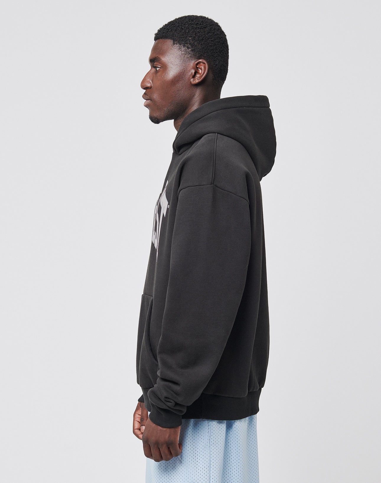 A man stands in profile against a plain background, wearing a LIVE FAST DIE YOUNG Live Fast College Hooded sweatshirt in black with the hood down and light blue mesh shorts. His arms are at his sides as he looks straight ahead.