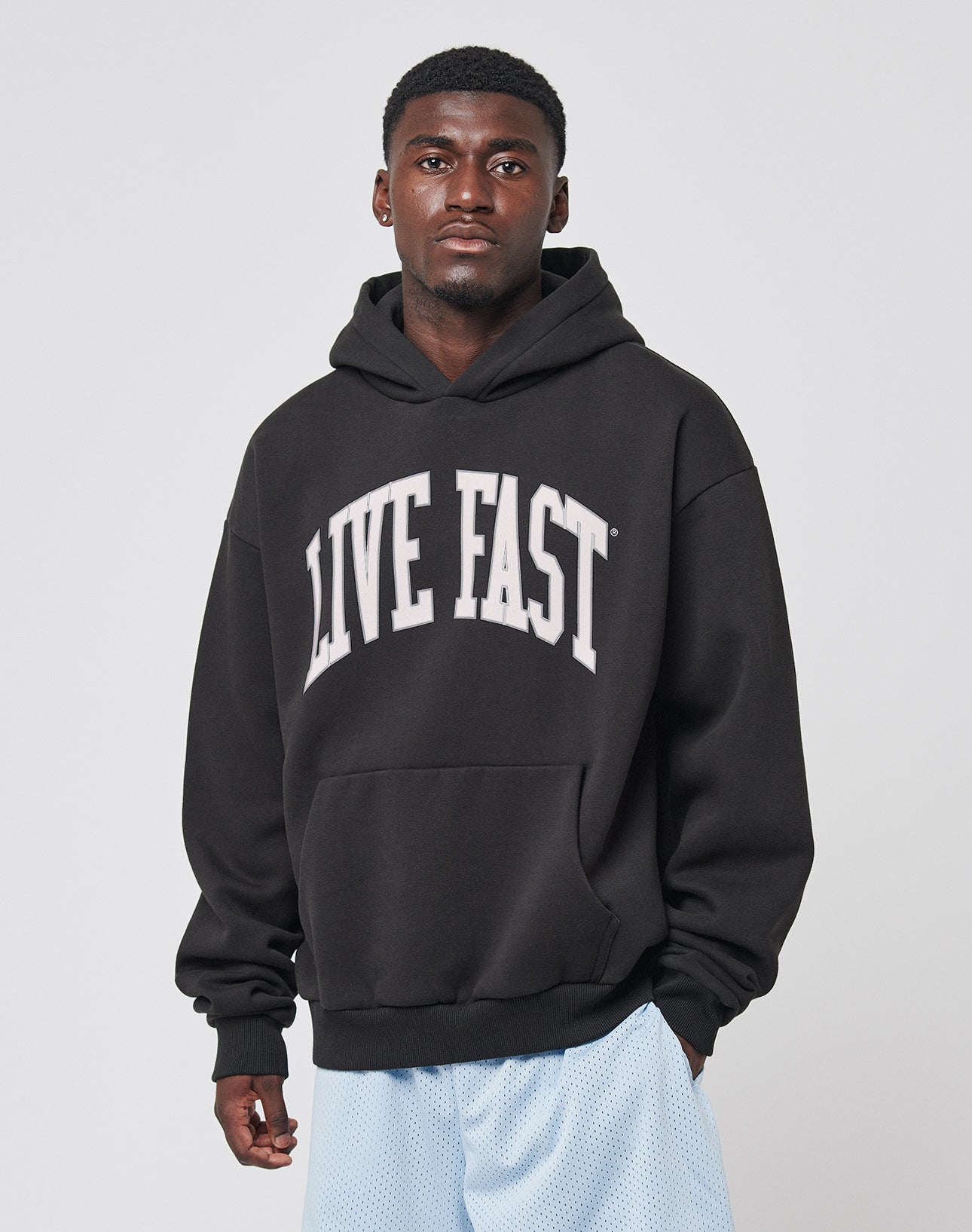 A man wearing the LIVE FAST DIE YOUNG Live Fast College Hooded in black, featuring a bold white LIVE FAST front print and an oversize fit in soft Baumwolle, stands against a light background, paired with light blue shorts and a neutral expression.