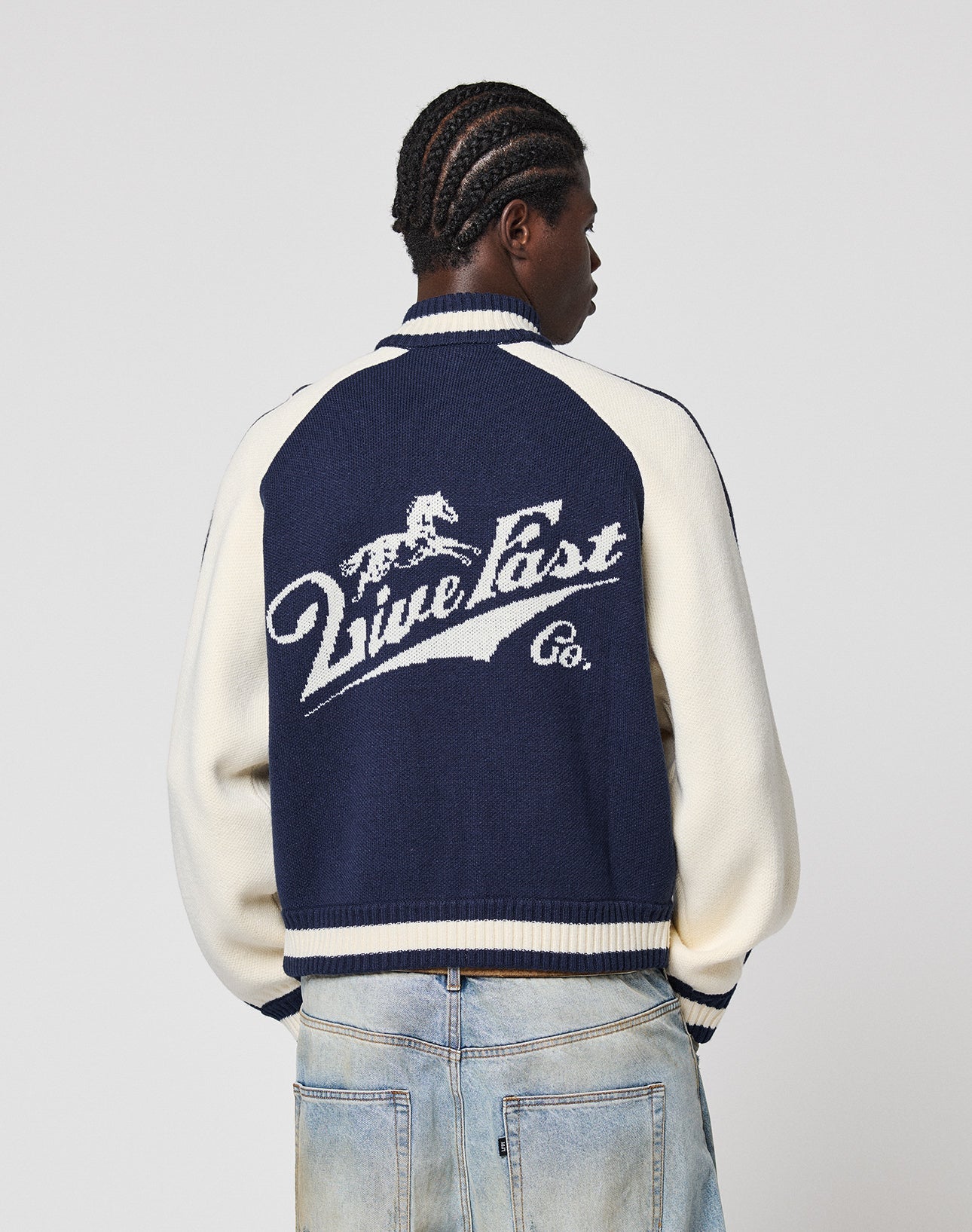 A person with short braids wears a Boxy Knit Zip Jacket by LIVE FAST DIE YOUNG—a navy and cream varsity style with a horse graphic and "Live Fast Co." on the back—paired with light blue jeans, standing facing away from the camera.