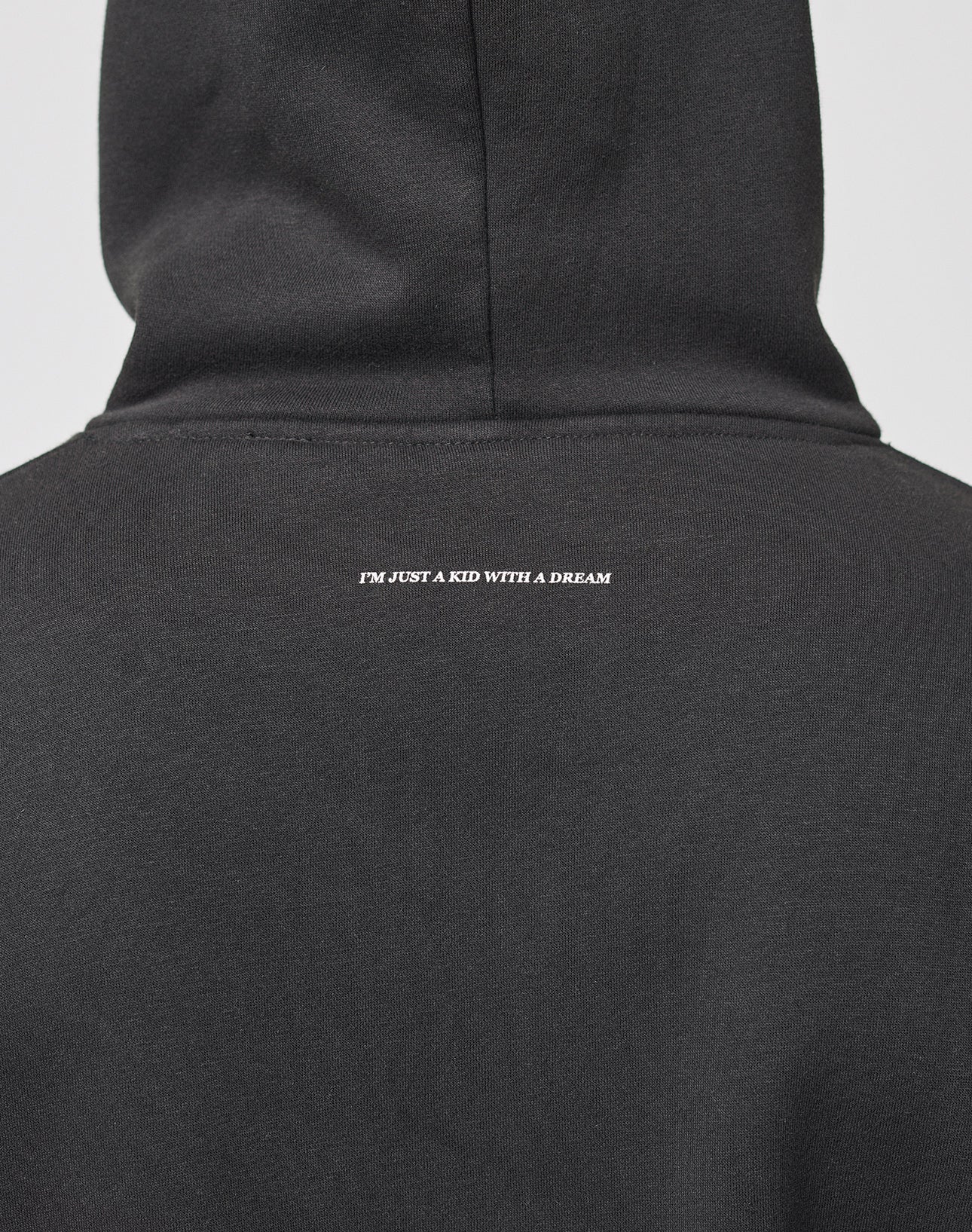 A person wearing a black Live Fast College Hooded by LIVE FAST DIE YOUNG in Oversize Fit, seen from the back, with IM JUST A KID WITH A DREAM printed in small white letters on the upper back.