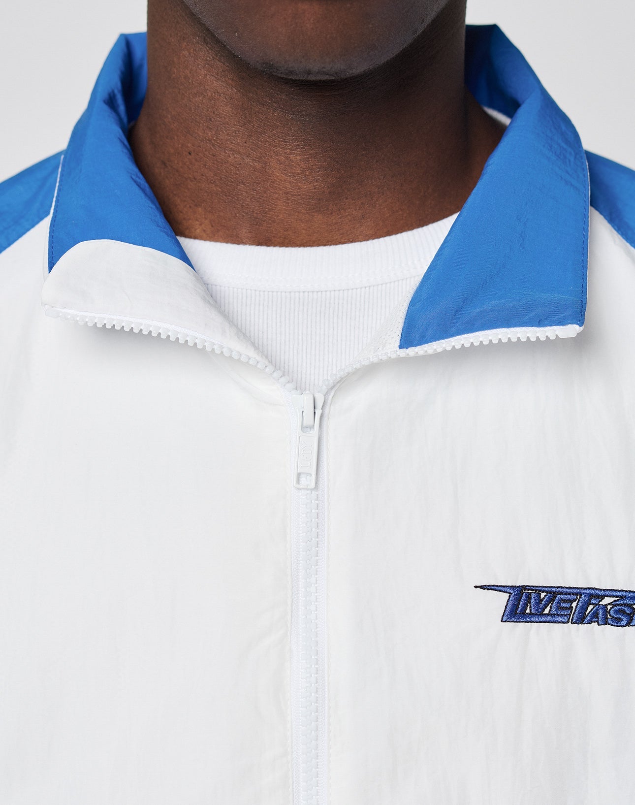 Close-up of a person wearing the unzipped LFDY Trackjacket 3000 by LIVE FAST DIE YOUNG, featuring a white polyamid body, blue collar, multilogo embroidery on the left, and branded zipper puller over a white shirt. Face is partially visible.