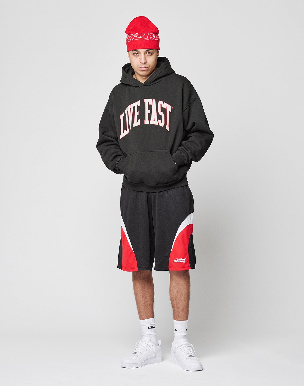 A person wearing the oversize fit Live Fast College Hooded by LIVE FAST DIE YOUNG, featuring a bold front print, pairs it with a red beanie, black and red shorts, white socks, and sneakers. The hoodie contains 65% Baumwolle. Hands are in the pocket.