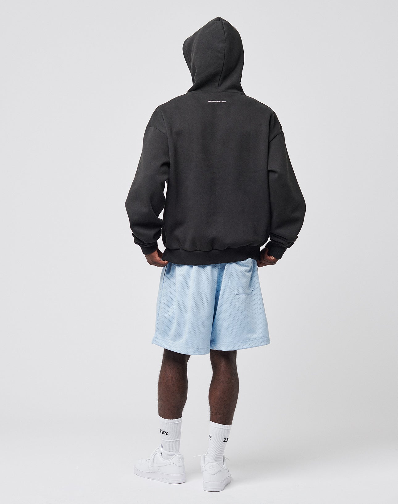 A person stands facing away against a plain white background, wearing the LIVE FAST DIE YOUNG Live Fast College Hooded in black (hood up), light blue athletic shorts, white socks, and white sneakers.