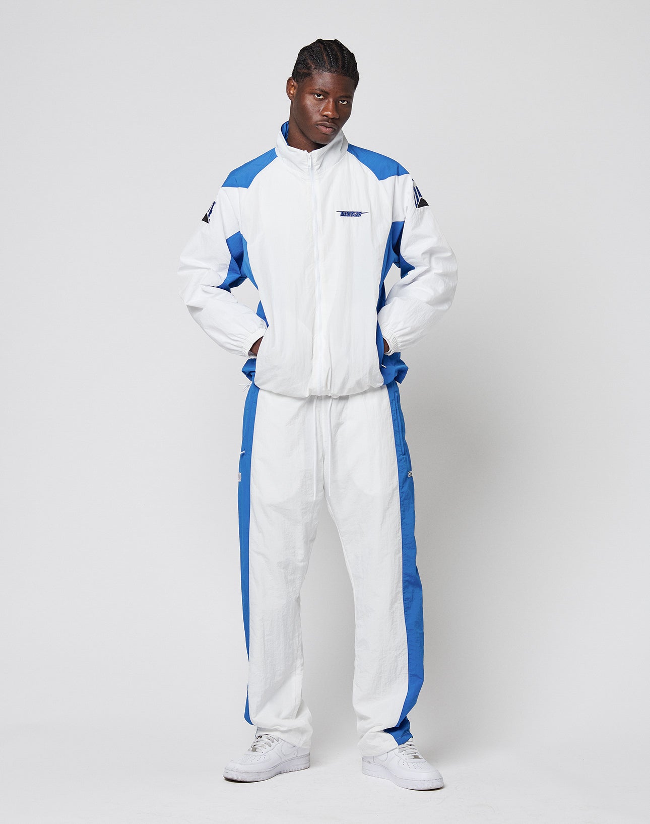 A person stands against a light background wearing LIVE FAST DIE YOUNGs LFDY Trackpants 3000 in white and blue with a wide fit and elastic waistband, paired with white sneakers, hands in pockets, looking at the camera neutrally.