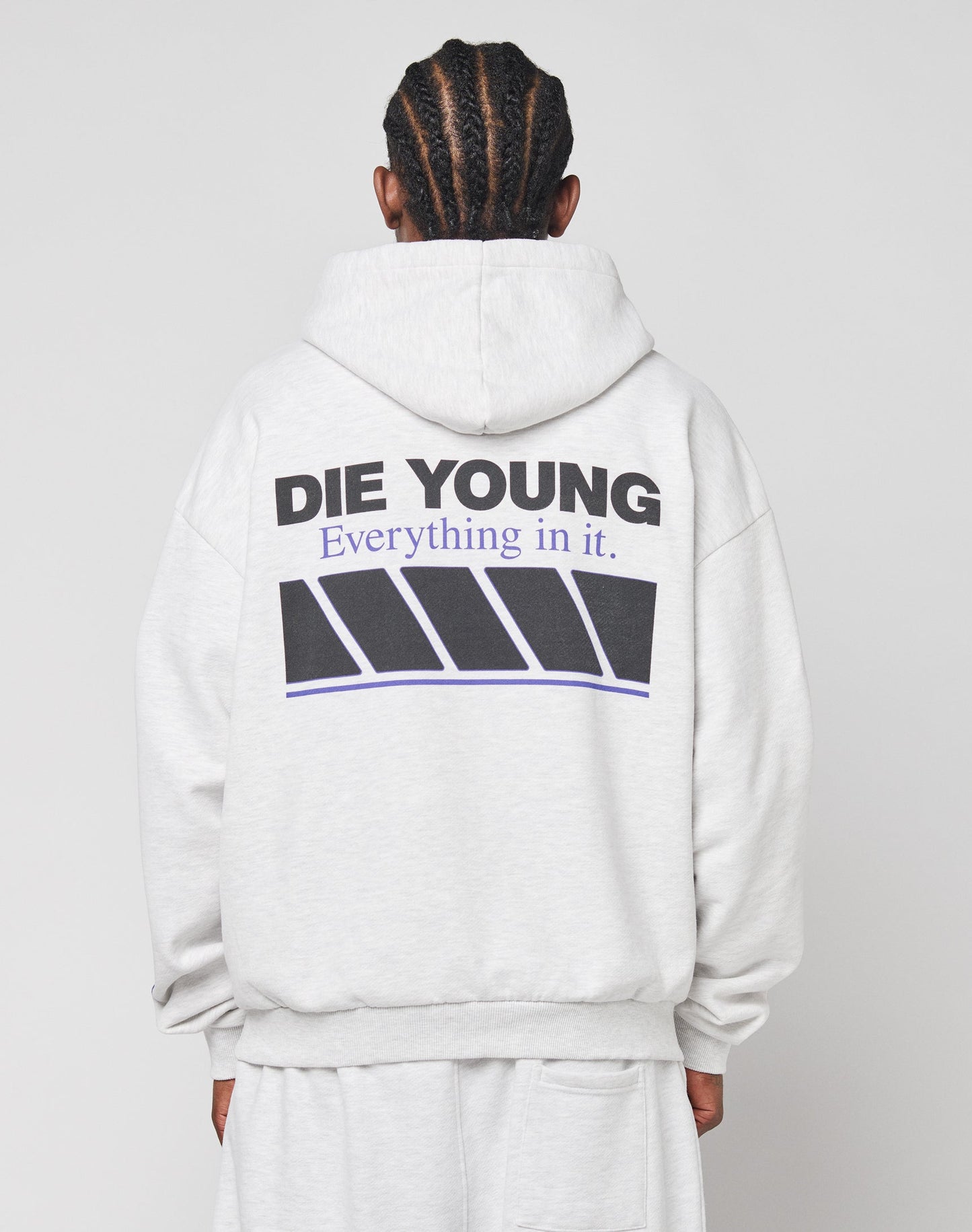 Person with braided hair seen from the back, wearing the oversize fit LFDY Racing Hooded by LIVE FAST DIE YOUNG in light gray Baumwolle Polyester. DIE YOUNG Everything in it. text sits above glossy black and purple angled rectangle graphics.