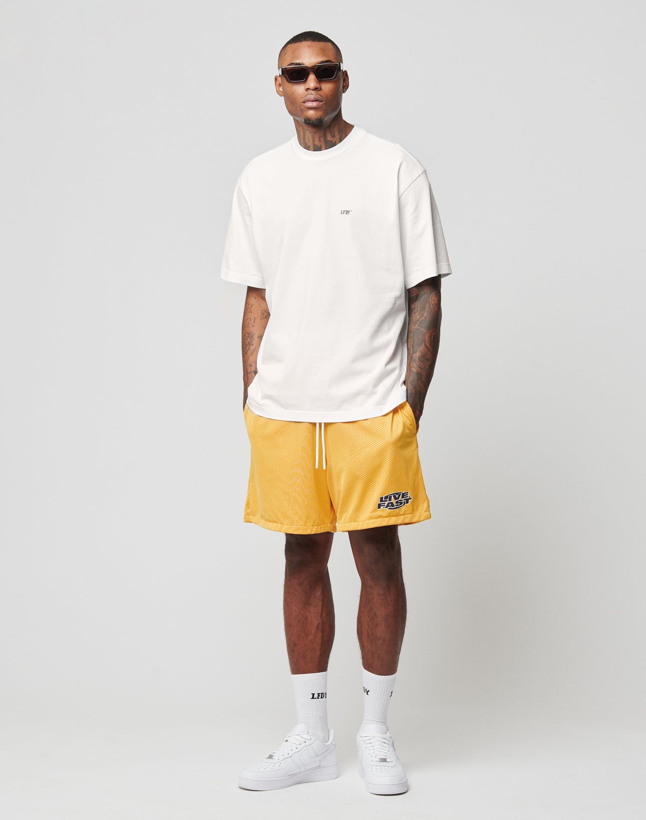 A man in sunglasses, a white t-shirt, LIVE FAST DIE YOUNG LFDY Oval Mesh Shorts with elastischer Bund, white crew socks, and white sneakers stands against a plain light gray background.