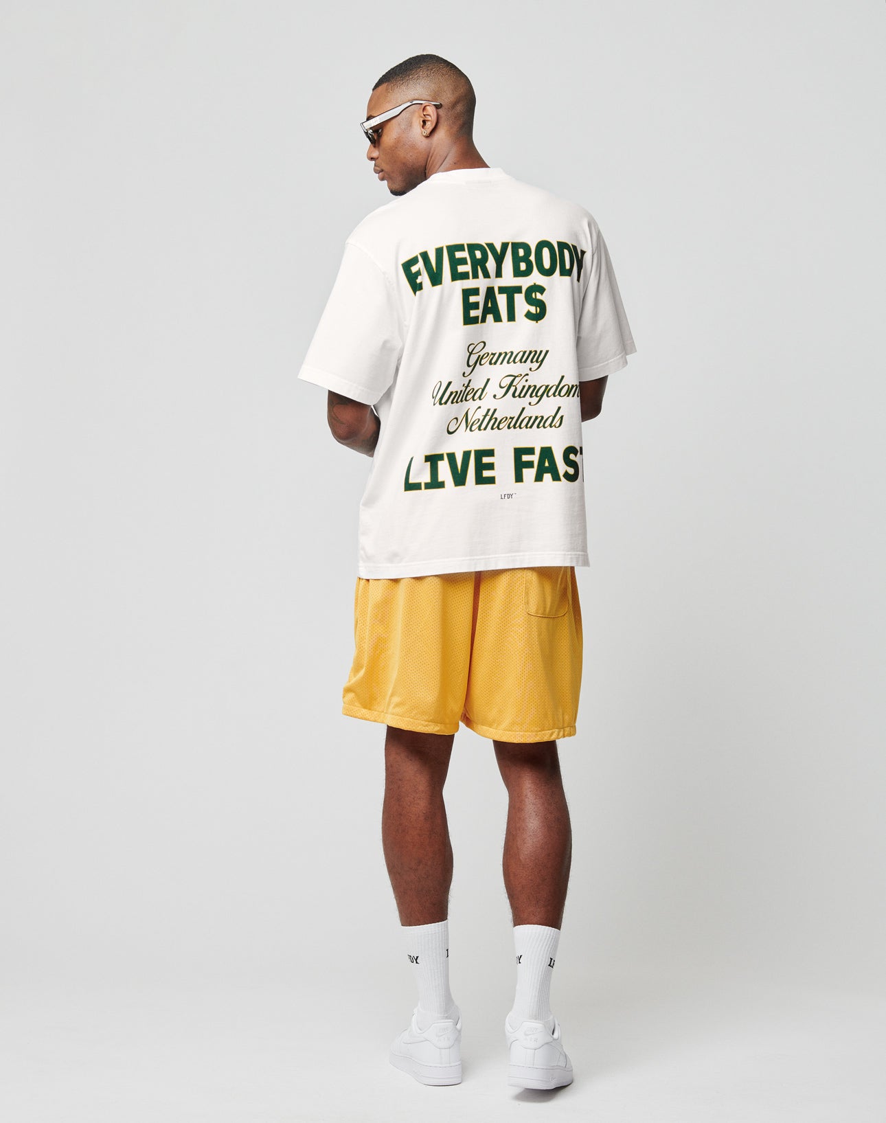 A man wearing sunglasses and the LIVE FAST DIE YOUNG Everybody Eats Tee—an Oversize Fit white T-shirt with green and yellow Backprint made from 100% Baumwolle—plus yellow shorts, white socks, and sneakers stands facing away from the camera.