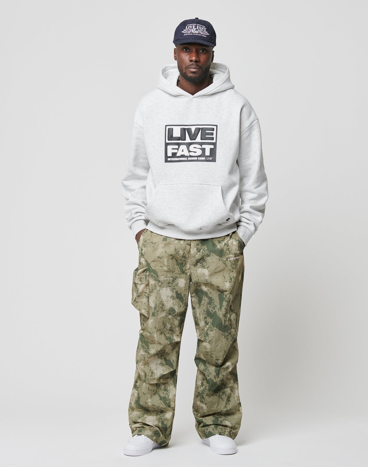 A man wearing a gray Live Fast Box Hooded sweatshirt by LIVE FAST DIE YOUNG, camouflage cargo pants, white sneakers, and a navy cap stands against a plain white background with his hands in his pockets.