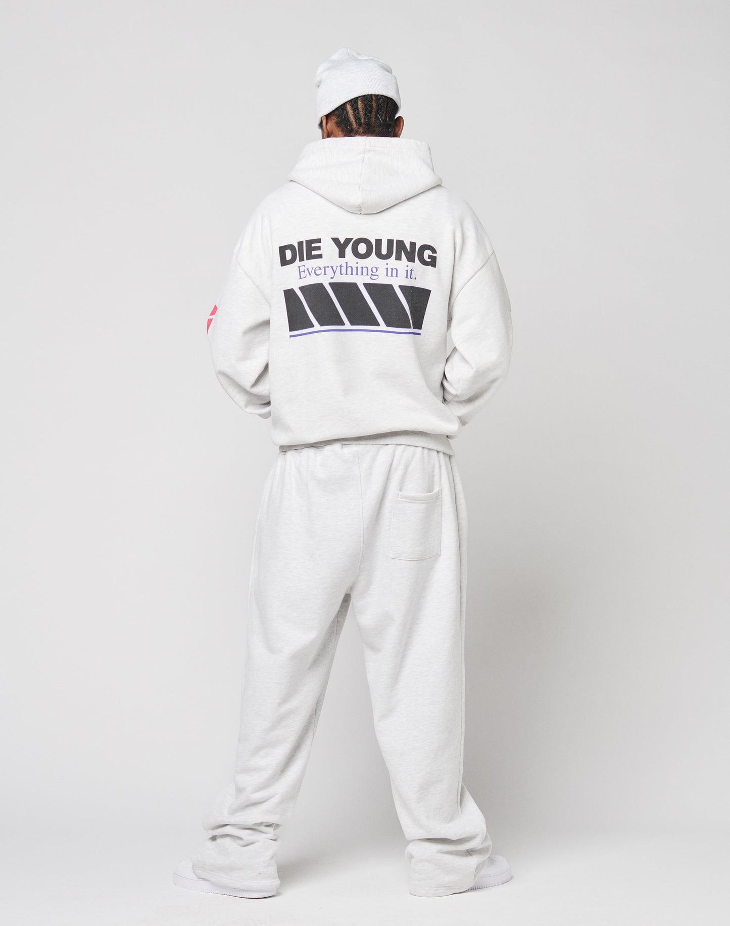 A person stands facing away, wearing an oversize fit light gray LFDY Racing Hooded and sweatpants from LIVE FAST DIE YOUNG. The hoodie has bold black DIE YOUNG Everything in it. text, glossy print accents, and diagonal stripe graphics.