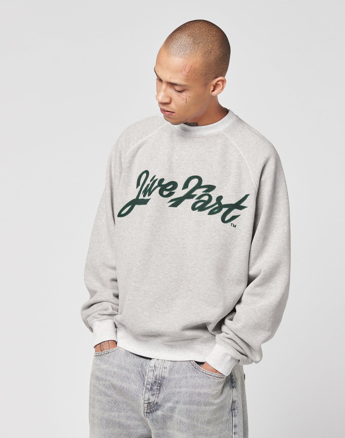 A person wears the LIVE FAST DIE YOUNG LFDY Script Sweater, a light gray oversized sweatshirt with raglan sleeves and Live Fast in dark green script on the front, looking down with hands in jean pockets against a plain background.
