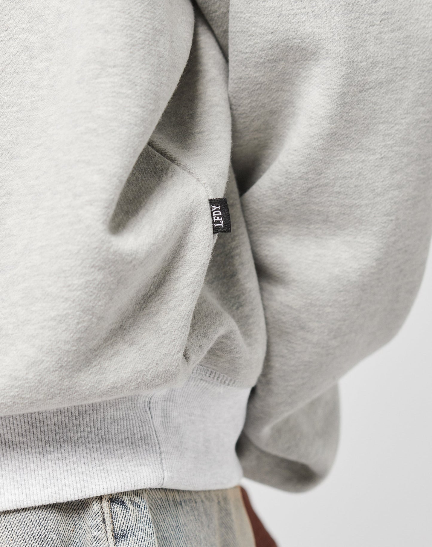 Close-up of a person wearing the LFDY Script Sweater by LIVE FAST DIE YOUNG in light grey with oversize fit, raglan sleeves, cotton-polyester blend, small black ACT tag on the side seam, paired with light blue denim jeans.