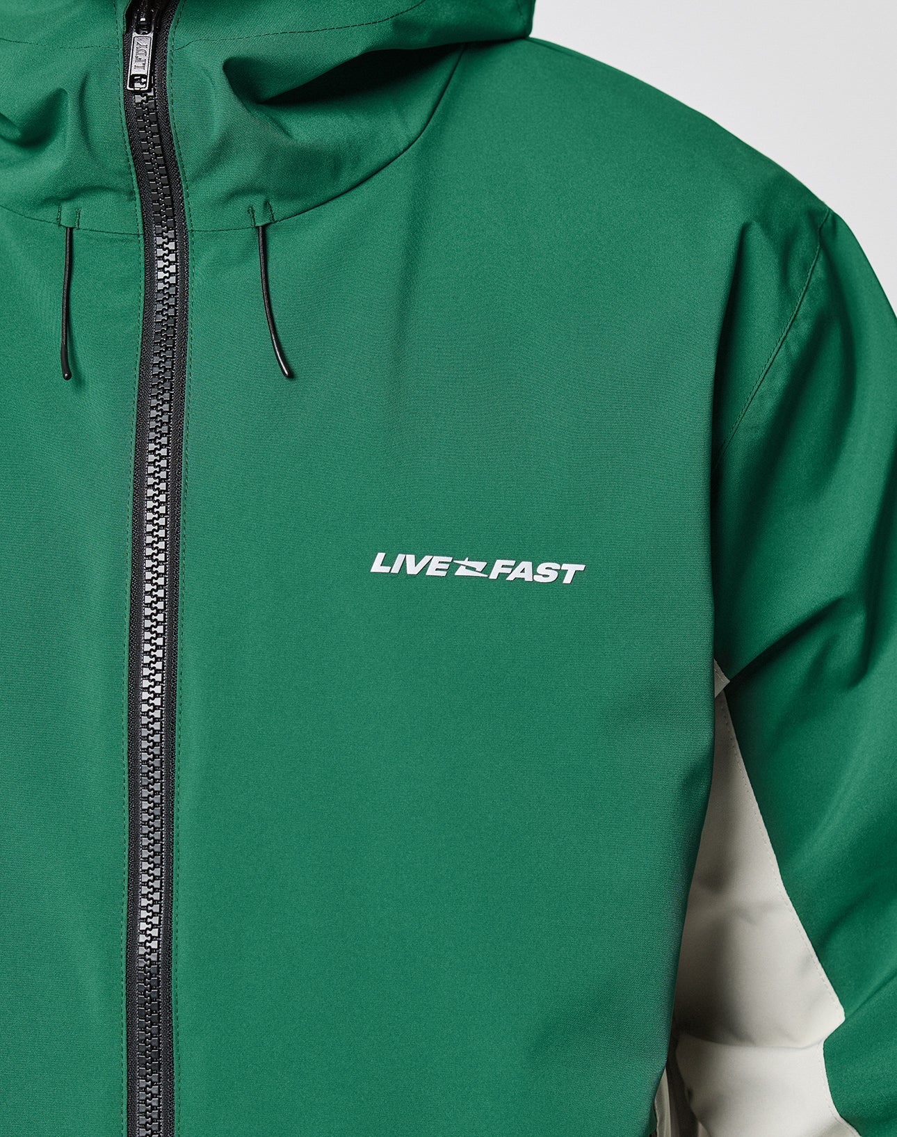 LIVE FAST DIE YOUNG Basic Shell Jacket with an oversize fit, black water-repellent zipper, hood, and white reflective LIVE FAST print on the left chest. Right sleeve in contrasting light beige. Only the upper torso of the jacket is visible.