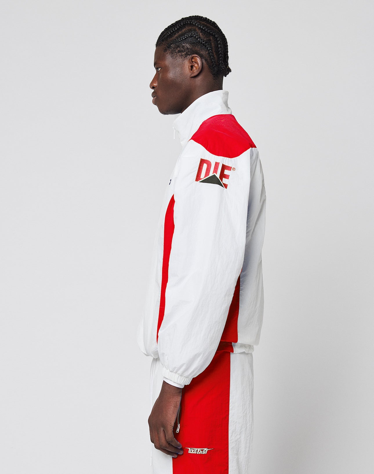 A person with braided hair stands in profile wearing the LFDY Trackjacket 3000 by LIVE FAST DIE YOUNG, a regular fit white and red tracksuit featuring bold DIE lettering on the sleeve and a branded zipper pull. The background is plain and light.