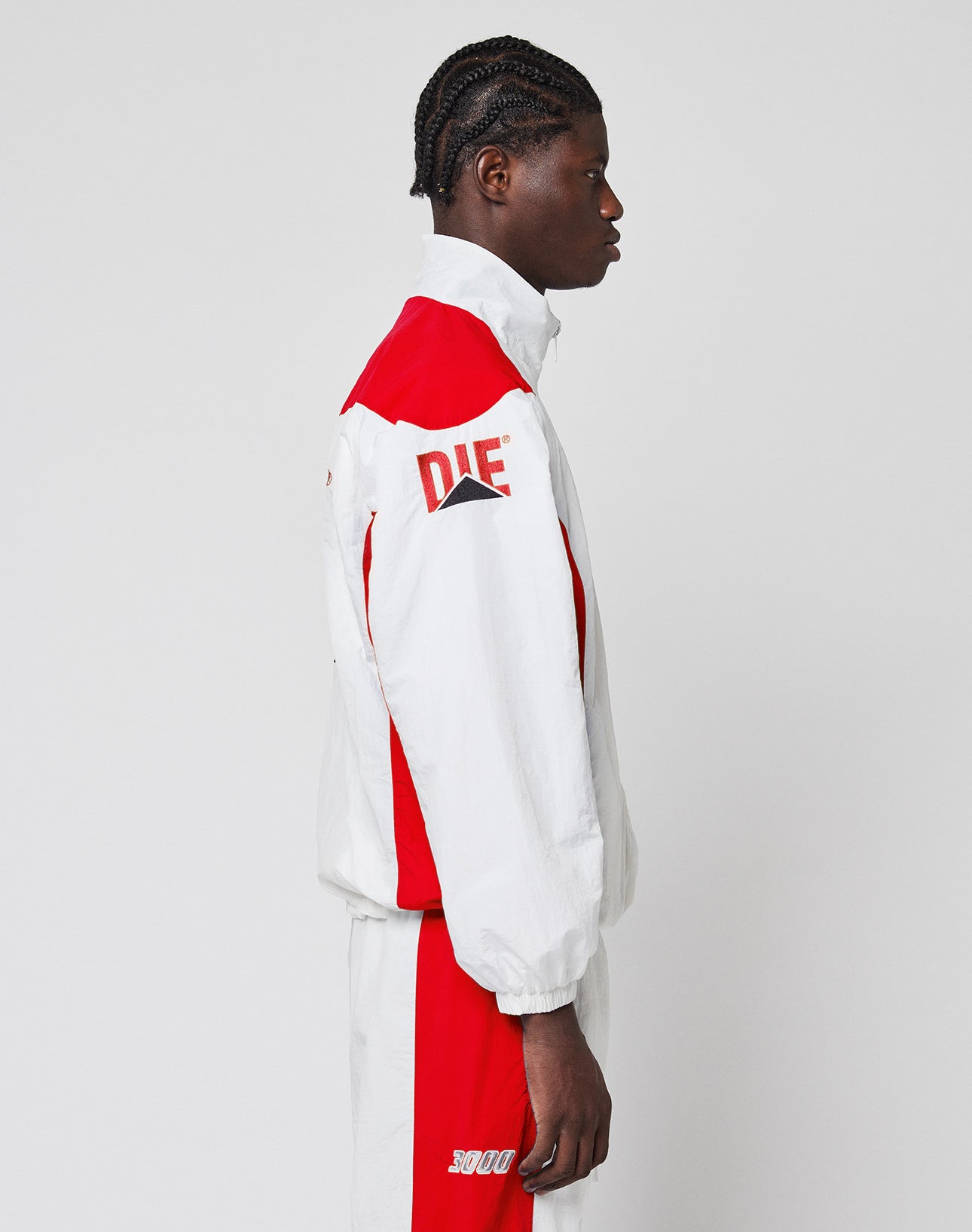 A person with braided hair stands in profile, wearing the LIVE FAST DIE YOUNG LFDY Trackjacket 3000—a regular-fit, color-block tracksuit with DIE on the sleeve, 5000 on the shorts, and an elastischer Kordelzug.