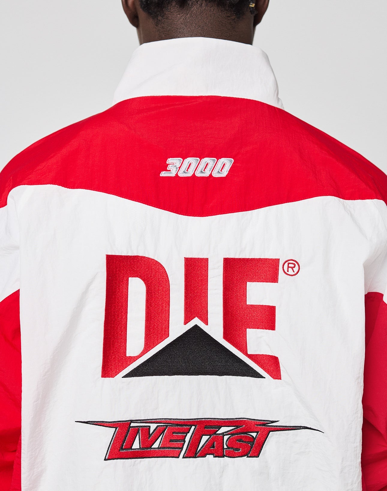 A person wearing the LFDY Trackjacket 3000 by LIVE FAST DIE YOUNG, a red and white jacket featuring 3000 embroidered at the top, Multilogo Stick DIE across the center, and LIVE FAST in stylized text at the bottom on the back.