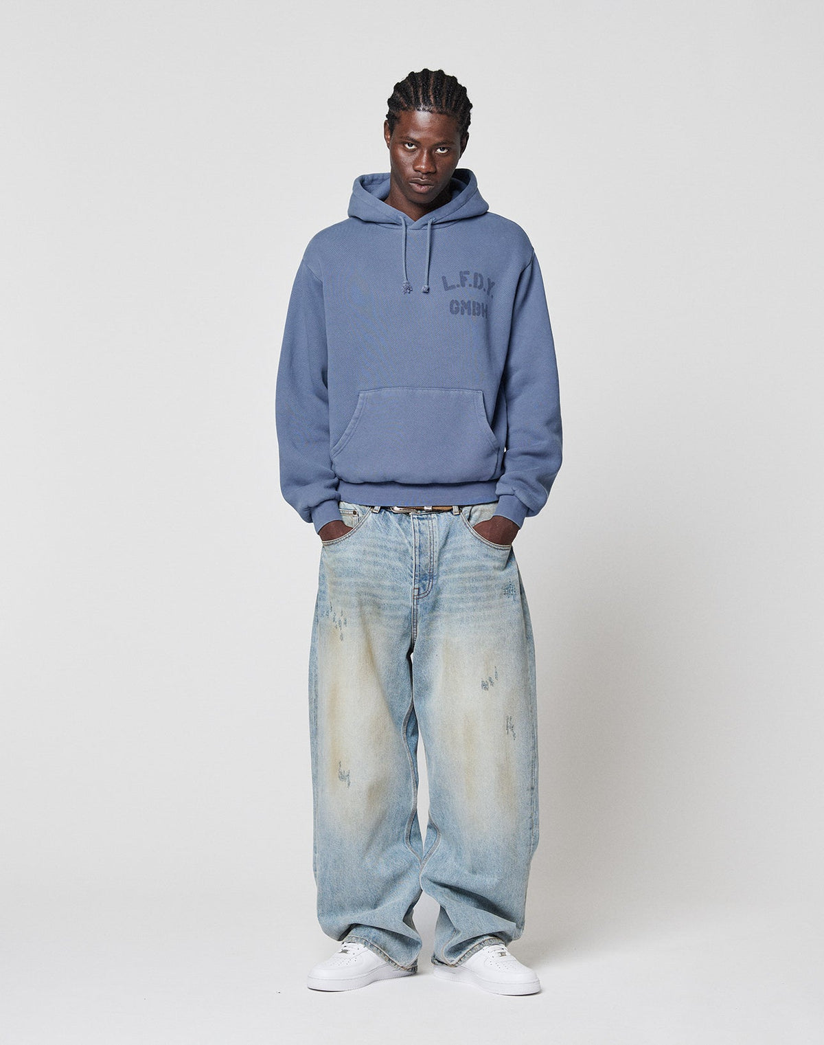 A person stands against a plain background wearing a blue LIVE FAST DIE YOUNG LFDY GmbH Hooded, loose-fitting faded jeans with Vintage Waschung, and white sneakers, hands in pockets with a neutral expression.