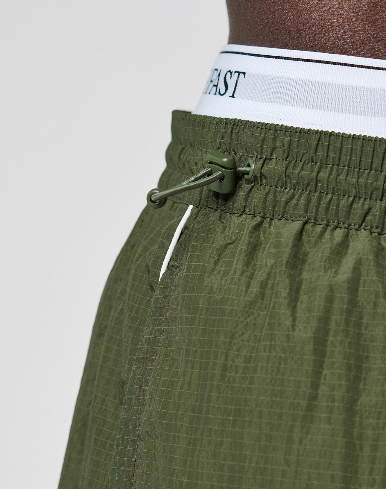 Close-up of a person wearing LFDY Nylon Trackshorts by LIVE FAST DIE YOUNG in olive green, showing a wide fit, adjustable drawstring, textured polyamide fabric, and a white logo-print waistband with black text.