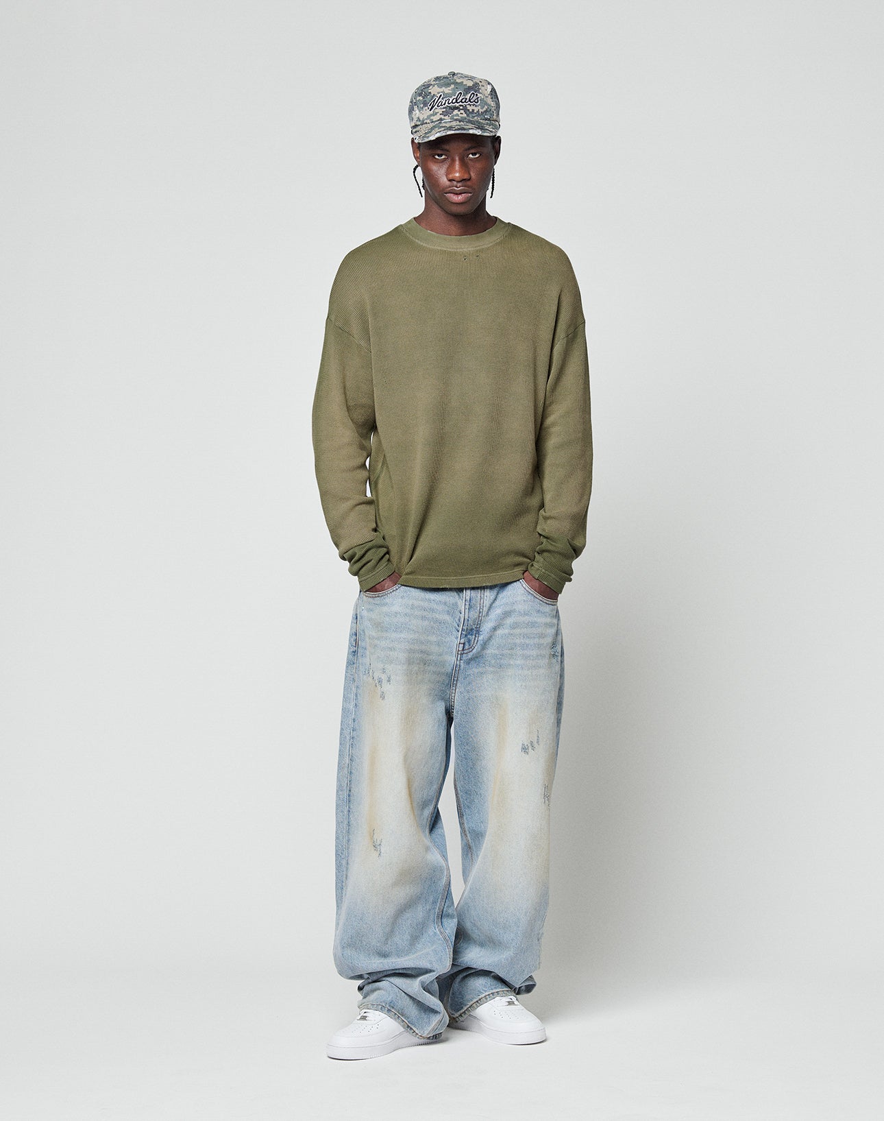 A person faces forward with hands in pockets, wearing a green LFDY Rib Knit Longsleeve by LIVE FAST DIE YOUNG, light blue baggy jeans with individuellem Wasch-Effekt, white sneakers, and a patterned cap against a plain background.