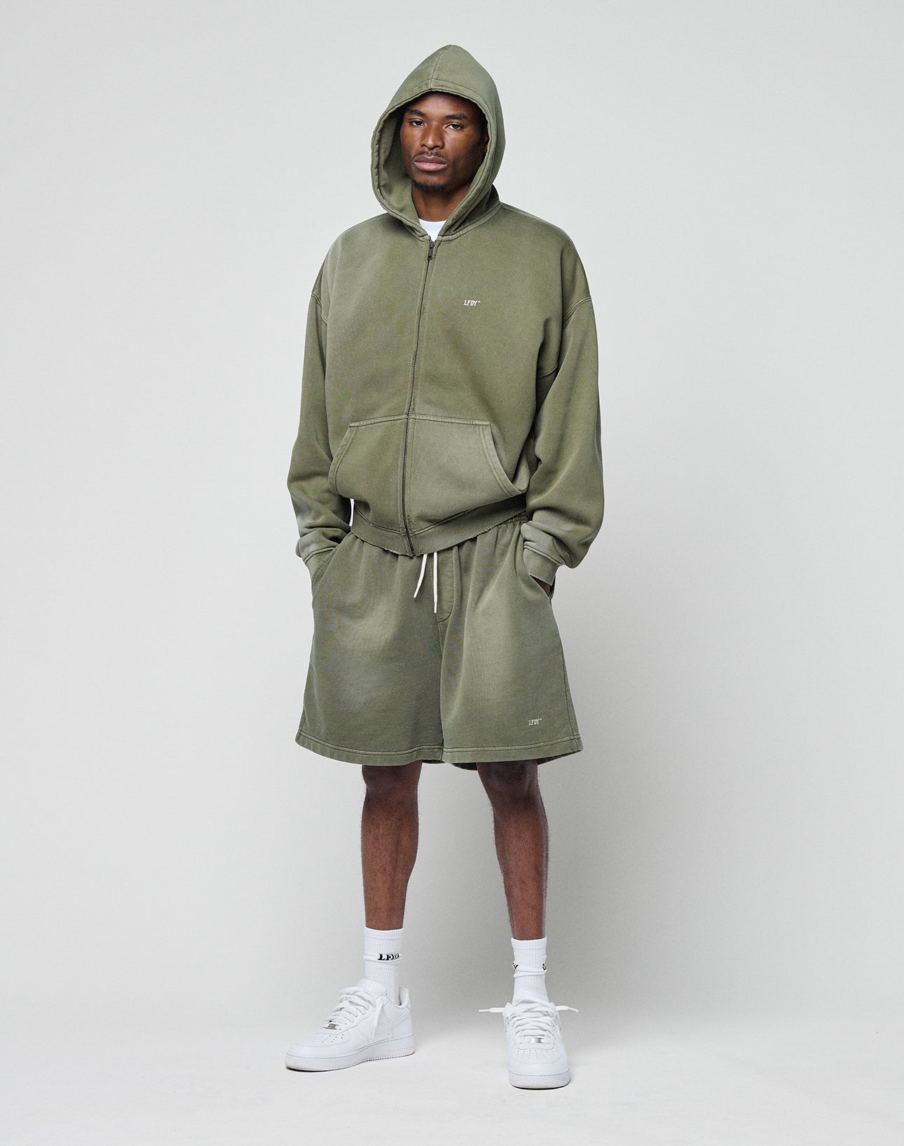 A man stands against a plain background wearing the LIVE FAST DIE YOUNG Basic Hooded Zip in olive green with a faded look, paired with matching baggy shorts, white socks, and white sneakers. His hands are in his hoodie pockets.