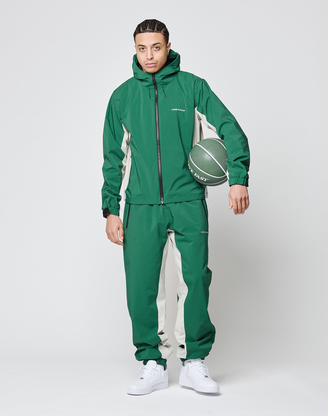 A man wears the LIVE FAST DIE YOUNG Basic Shell Jacket in an oversize green and beige fit with a wasserabweisender Reißverschluss, holding a green basketball under one arm. He faces forward against a plain light background in white sneakers.