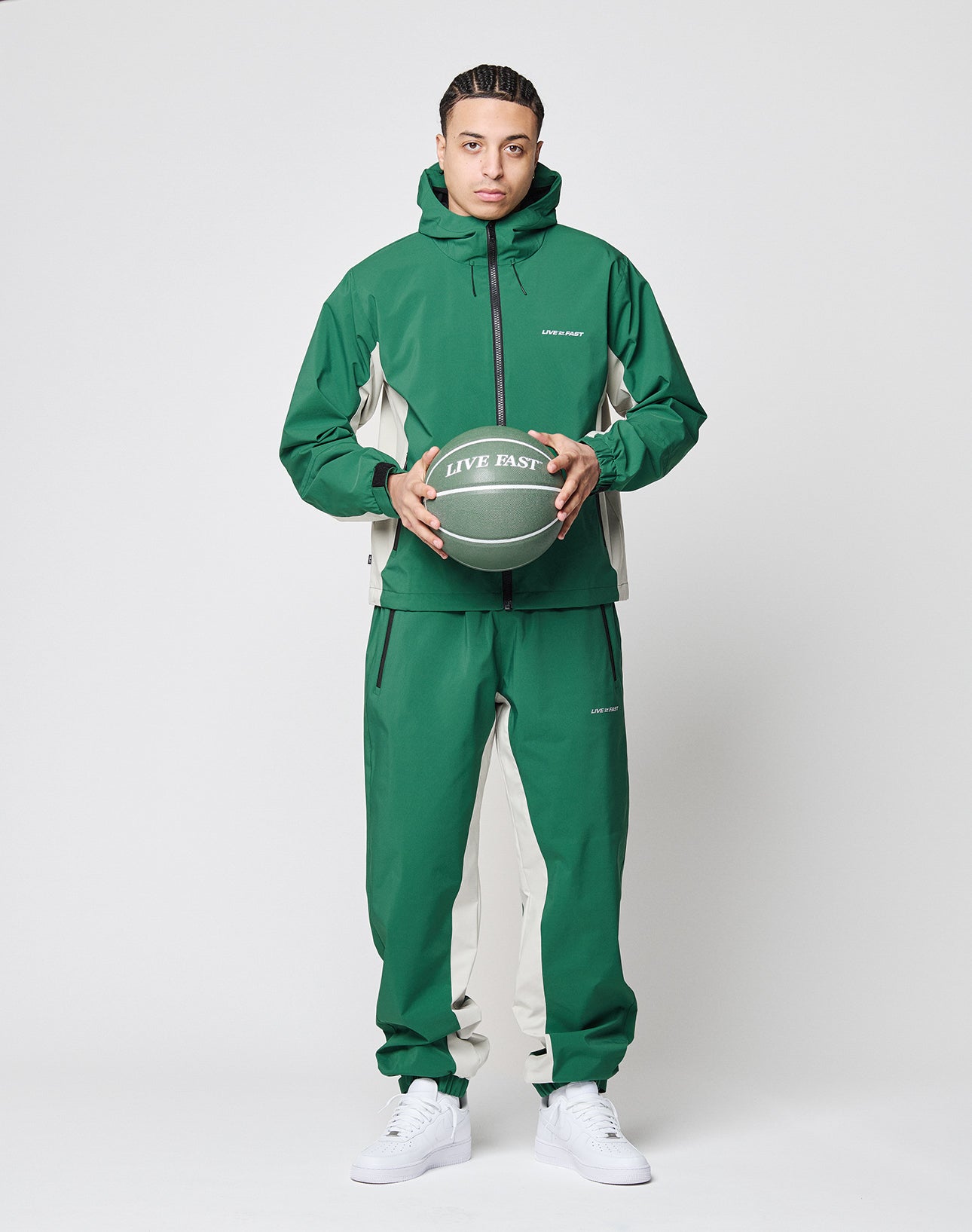 A person wears LIVE FAST DIE YOUNG Basic Shell Pants in an oversize green and beige fit with elastic waistband and drawstring, standing against a plain background, holding a green LIVE FAST basketball, wearing white sneakers, looking at the camera.