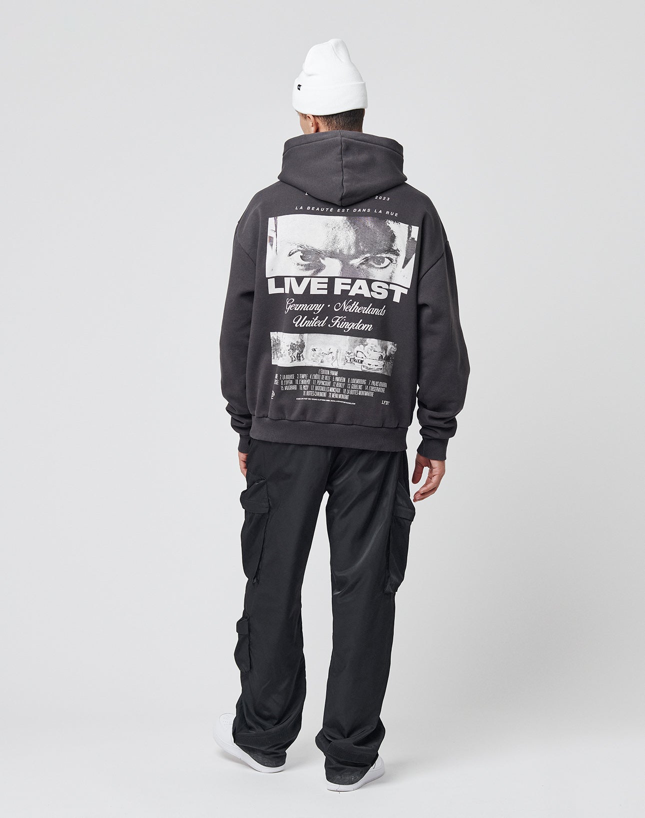 A person stands facing away, wearing a white beanie, a LIVE FAST DIE YOUNG LFDY Cinema Hooded Oversize Fit black hoodie with “LIVE FAST” on the back, black cargo pants, and white sneakers against a plain light background.