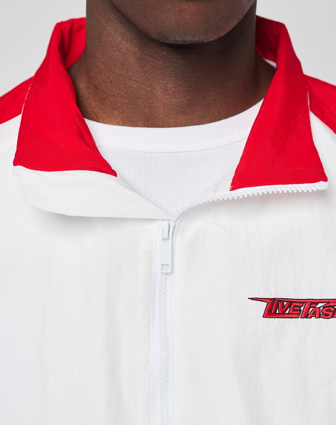 A person wearing the LIVE FAST DIE YOUNG LFDY Trackjacket 3000, a regular fit white zip-up with branded puller, red collar and shoulders, and METS embroidery over a ribbed white shirt; only their lower face and upper torso are visible.