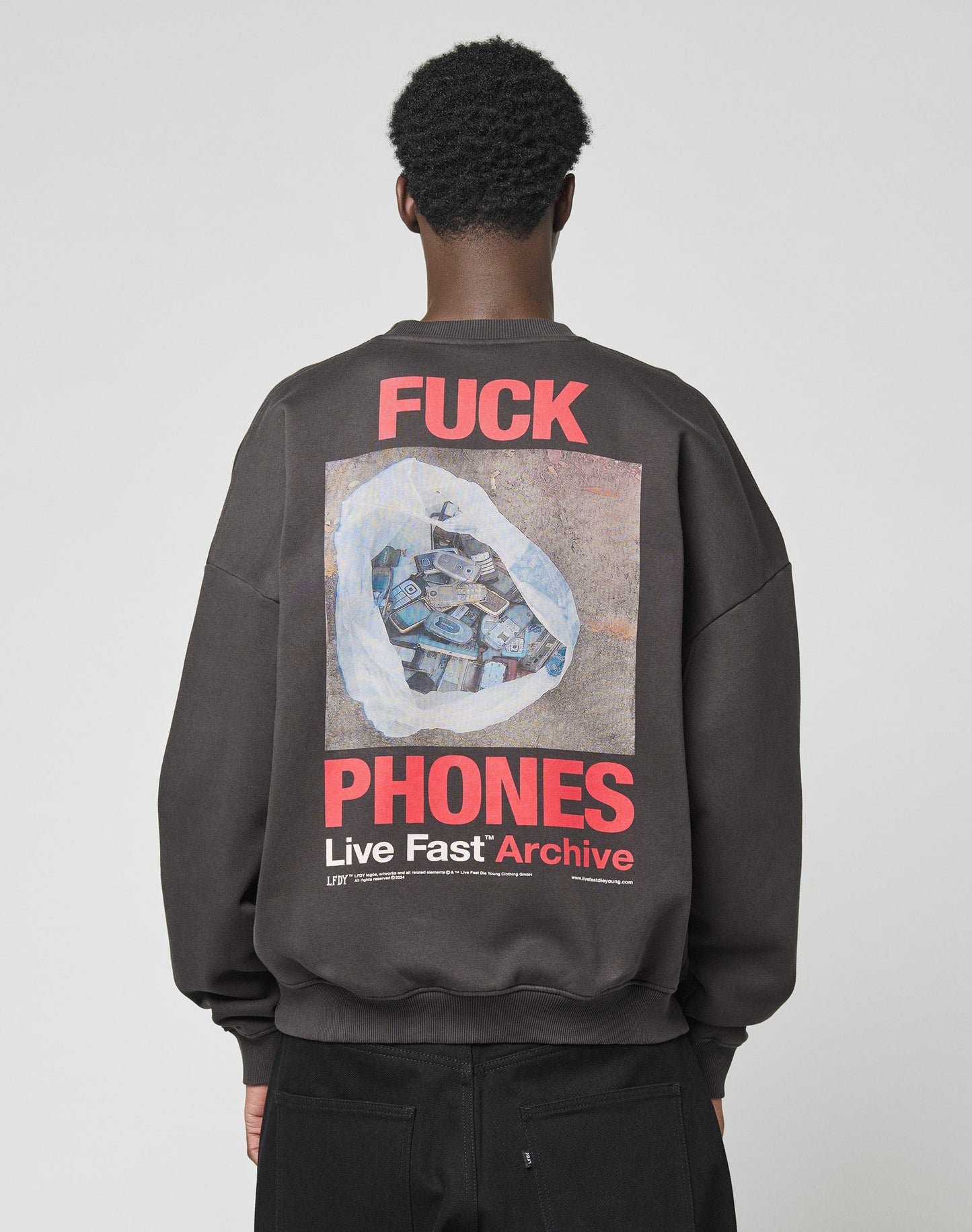 A person is seen from behind in the LFDY F*ck Sweater by LIVE FAST DIE YOUNG—an oversized black sweatshirt with bold red and white F*CK PHONES Live Fast Archive text and a hand holding phones graphic. Made from cotton and polyester.