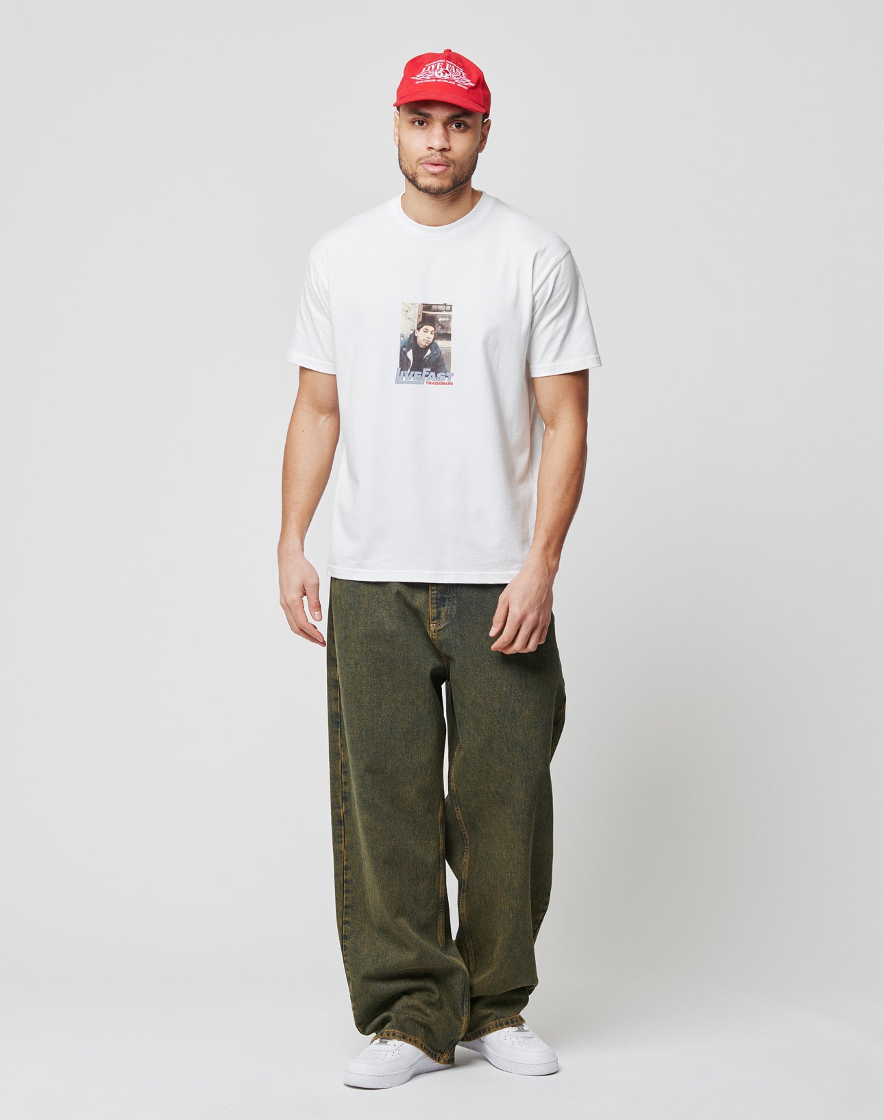 A man in a white graphic tee aus 100% Baumwolle, LFDY Baggy Denim von LIVE FAST DIE YOUNG im dunklen Grünton, weißen Sneakern und roter Baseballcap steht vor weißem Hintergrund und blickt nach vorn.
