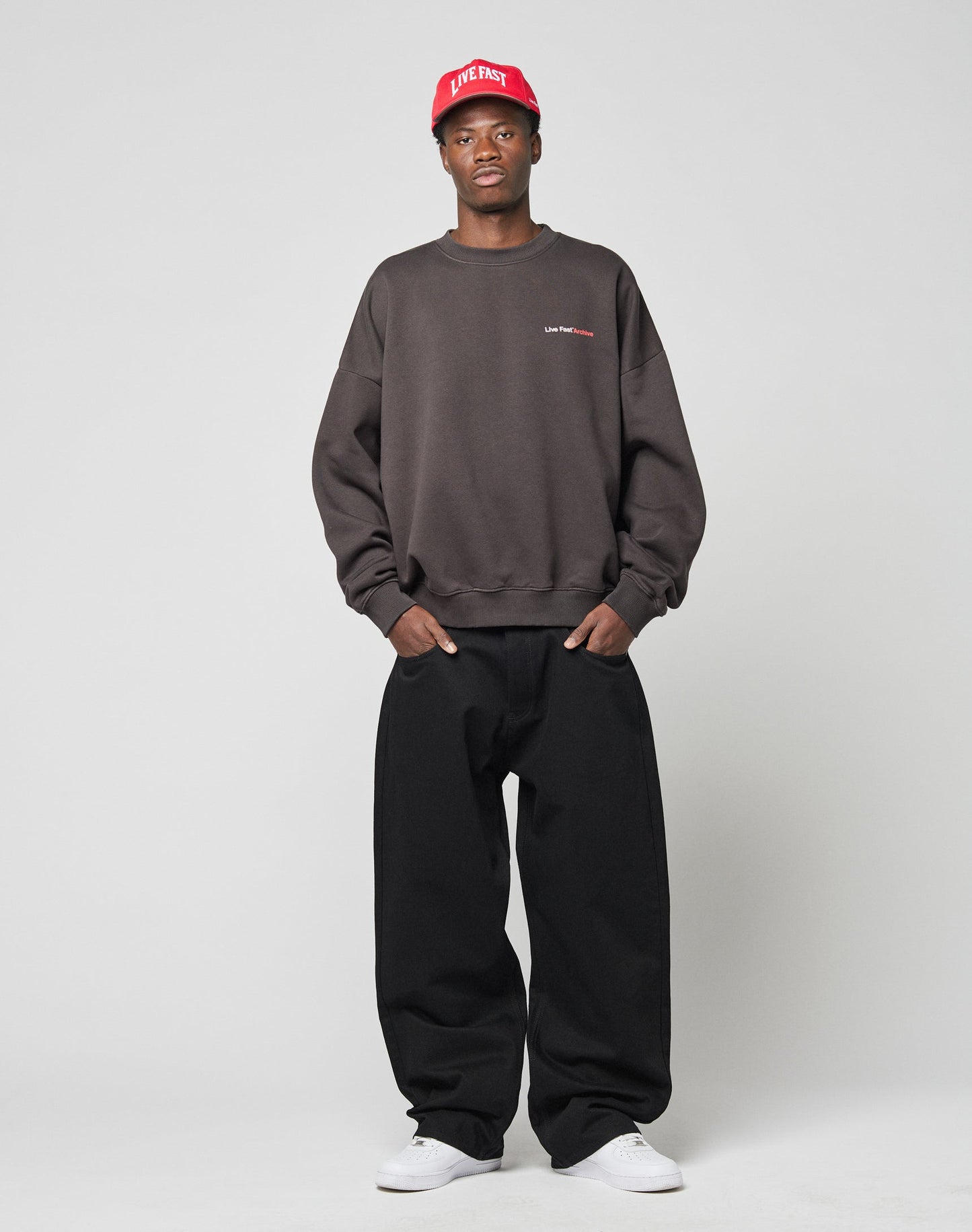 A person stands against a plain background wearing the LIVE FAST DIE YOUNG LFDY F*ck Sweater in an oversize fit, loose black pants, a red baseball cap, and white sneakers, with hands in pockets and a neutral expression.