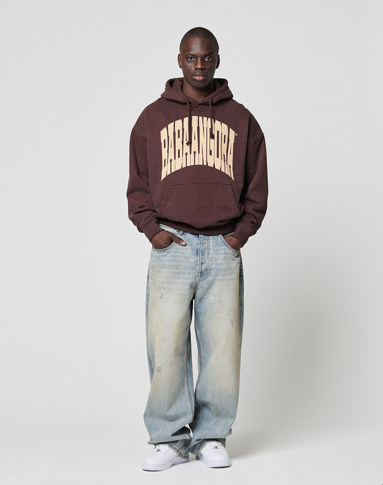 A person stands against a plain background wearing the LFDY x Baba Angora Hoodie by LIVE FAST DIE YOUNG, light blue baggy jeans, and white sneakers—a perfect streetwear look inspired by Berliner Stiftung style.