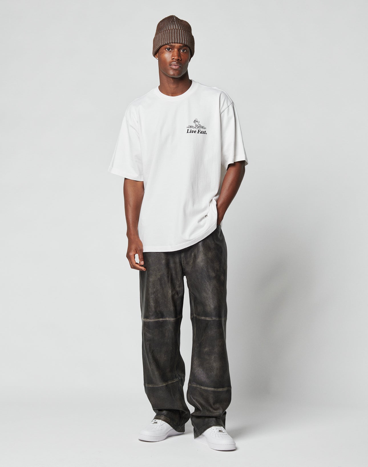 A man stands against a plain backdrop, wearing a brown knit beanie, loose white T-shirt with Flag Label detail, LIVE FAST DIE YOUNG Baggy Leather Pants, and white sneakers. One hand is in his pocket and he has a neutral expression.